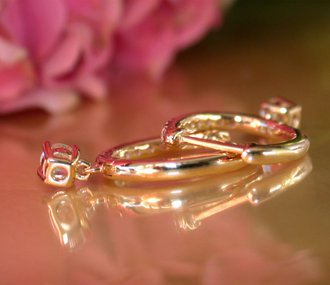 Goldene Creolen mit funkelnden Brillanten, Detailaufnahme auf glänzender Oberfläche, rosafarbener Hintergrund.