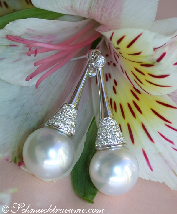 Detailansicht von Südseeperlen-Ohrsteckern mit Brillanten, verziert mit Diamantkette, vor weißem Blütenhintergrund.