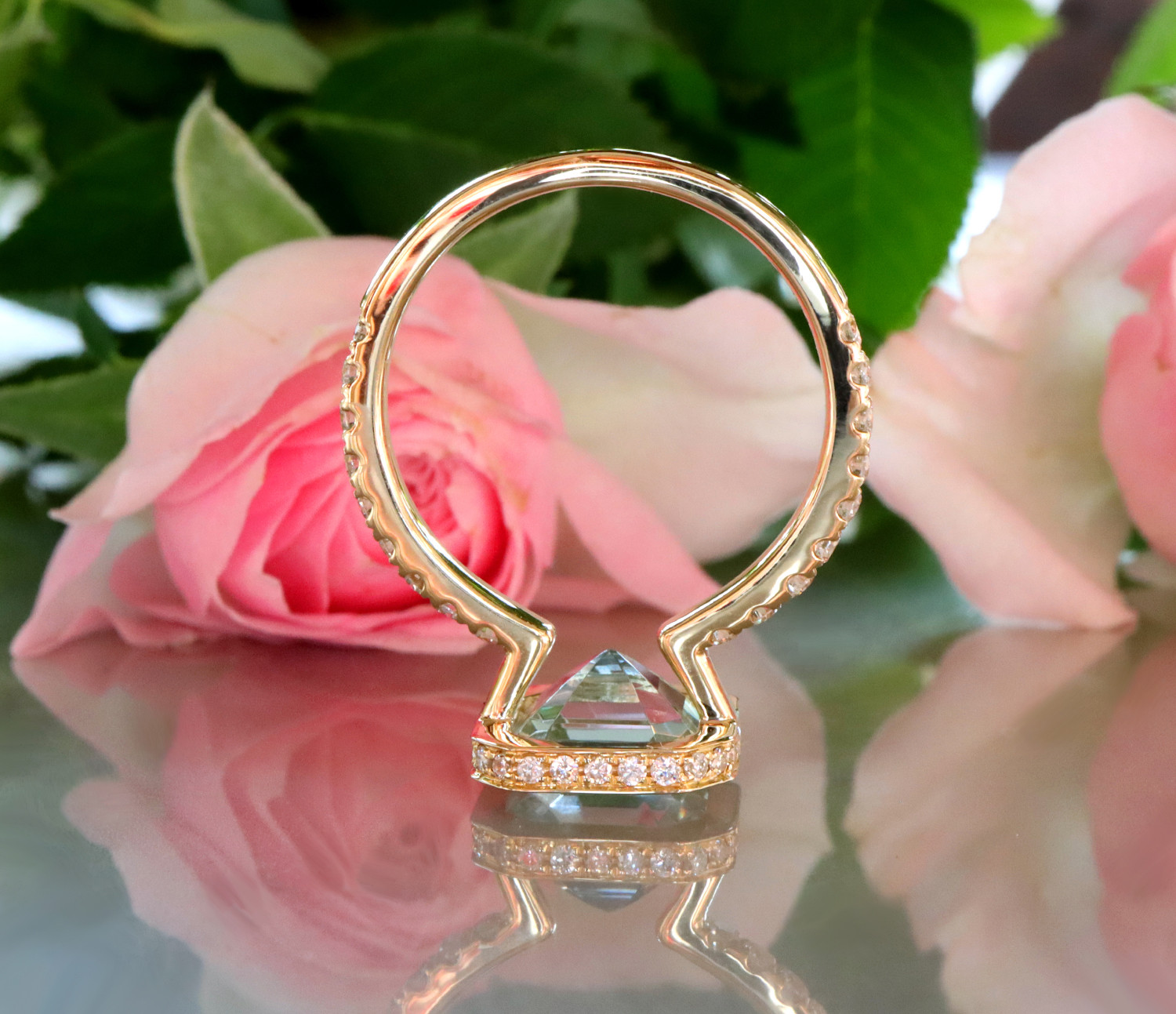 Goldener Aquamarin-Ring mit Diamanten auf glänzender Oberfläche, Rosenblüte im Hintergrund.