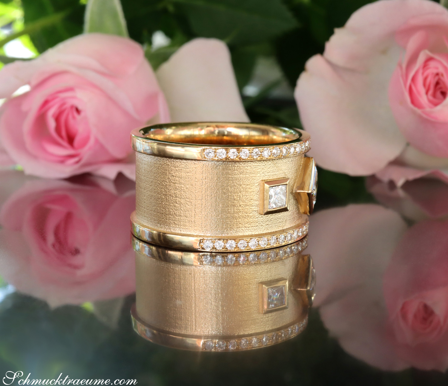 Goldener Diamantring mit Prinzessenschliffdiamanten und Diamantbesatz, Rosen im Hintergrund.