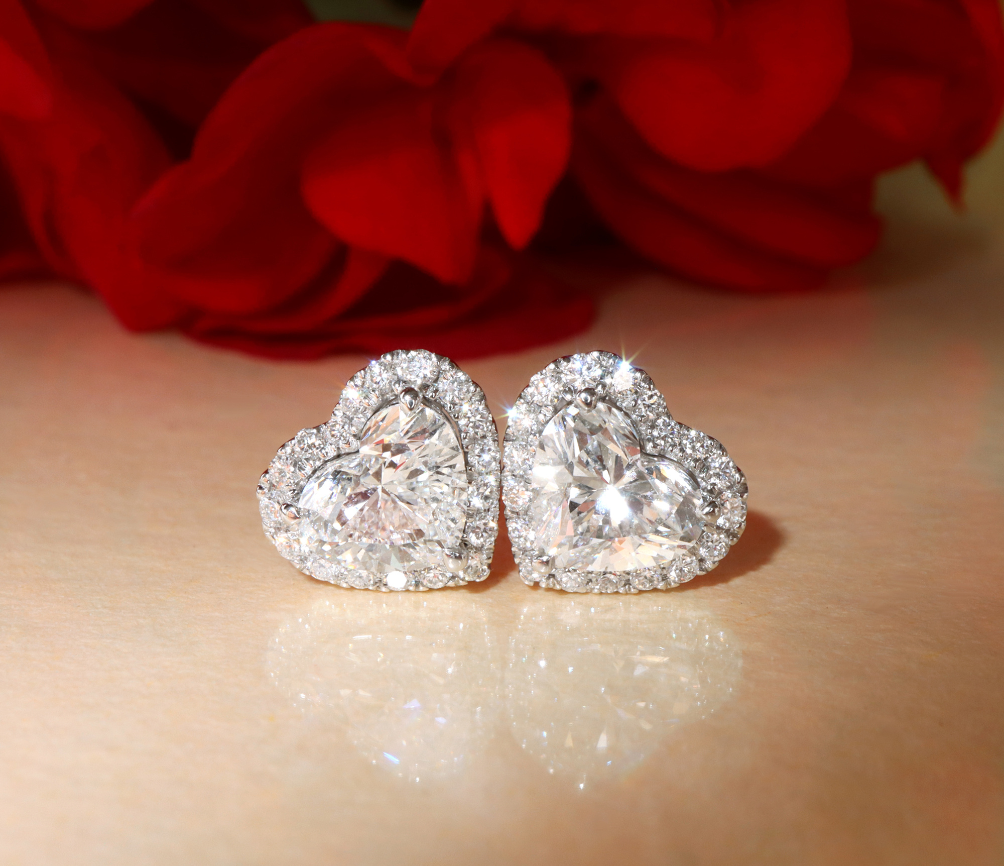 Herzformige Ohrstecker mit Diamanten, detailreich, in Weißgold 750, vor rotem Blütenhintergrund.
