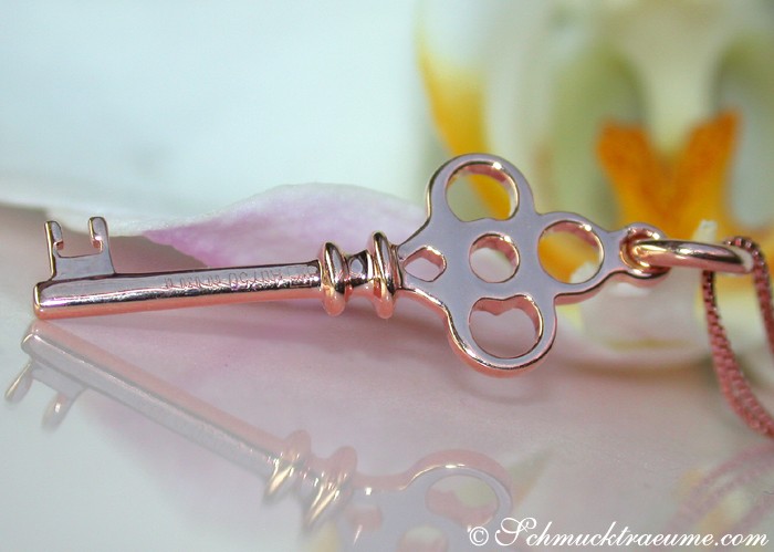 Detailaufnahme des femininen Diamanten-Schlüssel-Anhängers in Roségold, zarte Kette, Blumen im Hintergrund