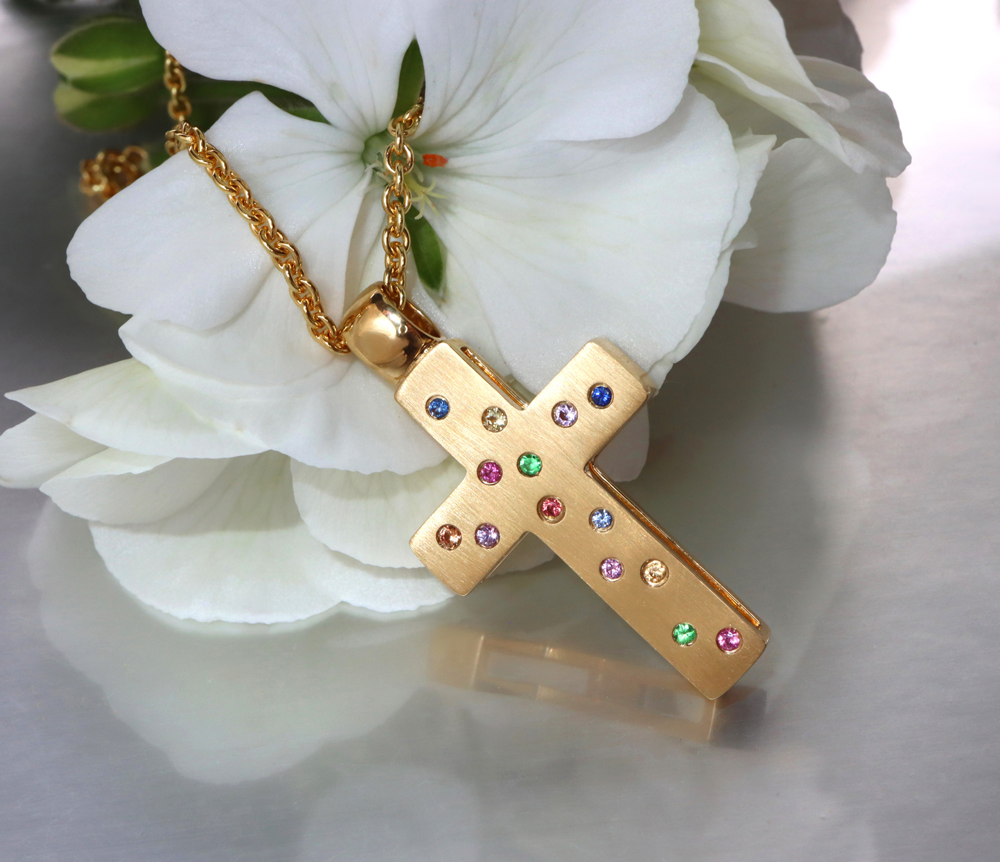 Goldene Kreuzanhänger mit Multicolor Saphiren und Brillanten, vor weißer Blume.