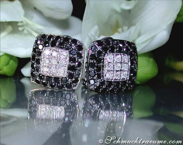 Schwarz-weiß Diamant-Ohrstecker im Carré-Design auf glänzender Oberfläche, Blüten im Hintergrund.