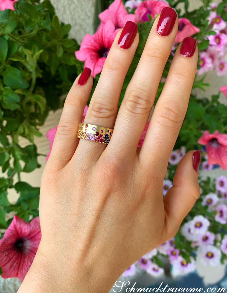 Gelbgold-Ring mit farbenfrohen Saphiren, Rubinen und Tsavorites, Nagellack rot, Blumen im Hintergrund