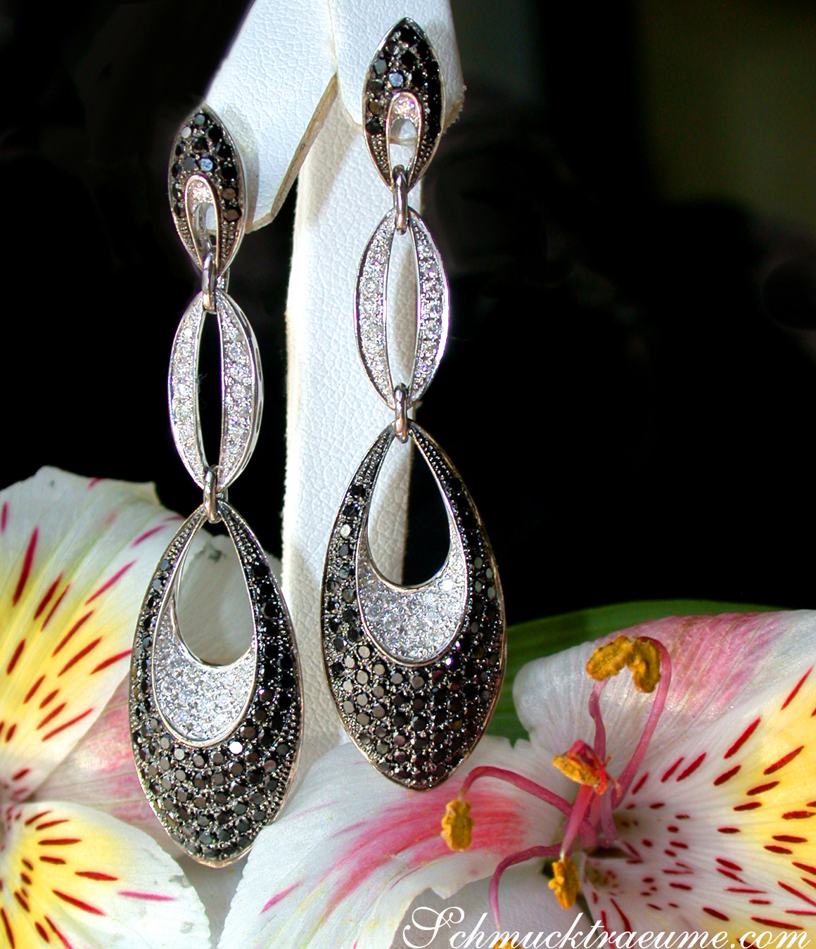 Weißgold-Ohrringe mit schwarzen & weißen Diamanten, elegante Formen, Blüten im Hintergrund.