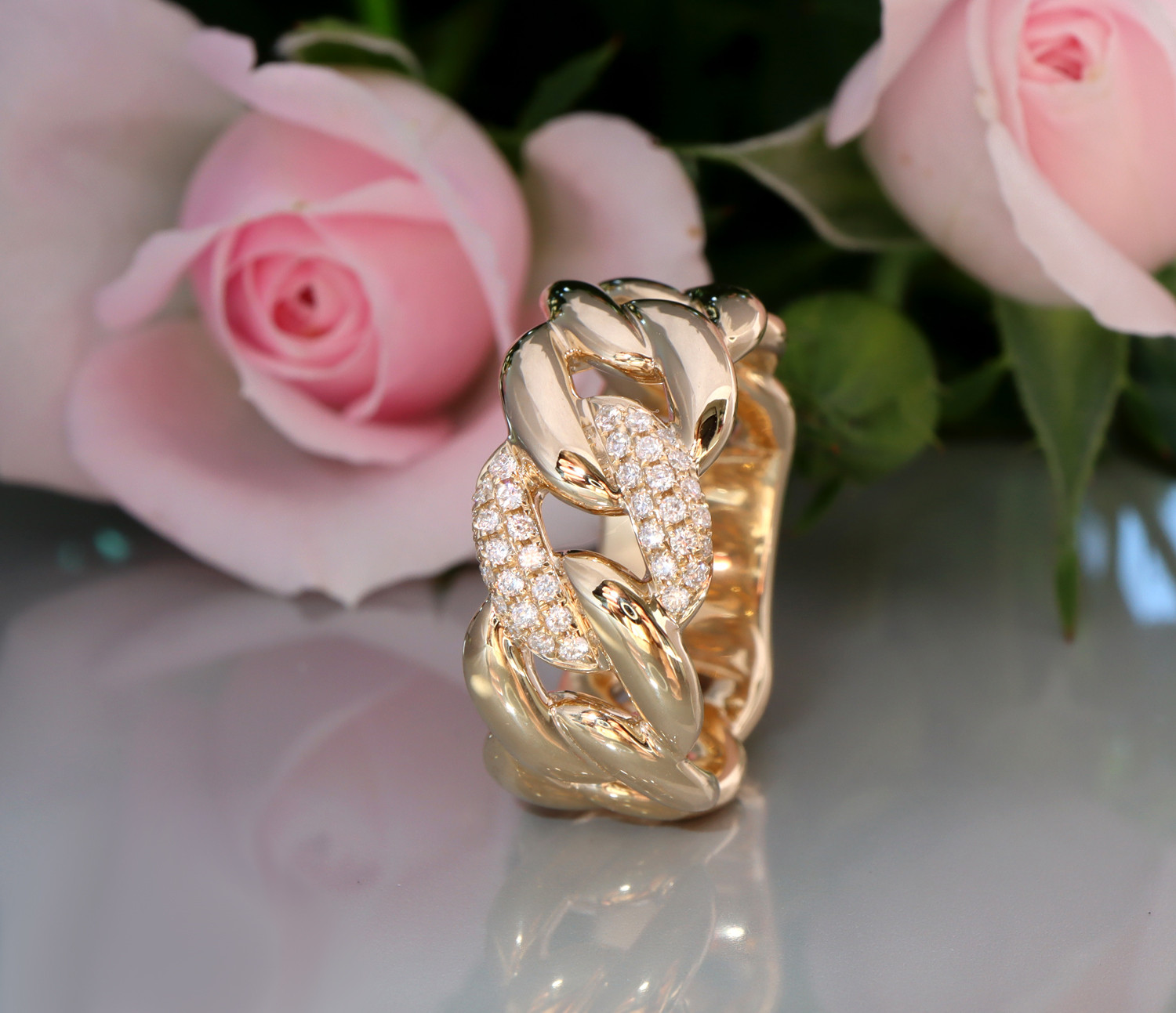 Goldener Panzerkettenring mit Brillanten, Detailaufnahme auf glänzendem Hintergrund, Rosen im Hintergrund.