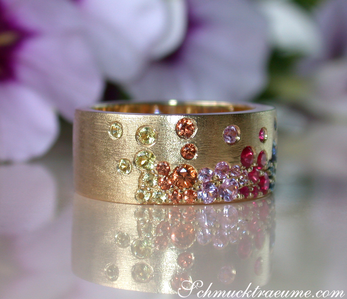 Goldener Ring mit farbenfrohen Saphiren, Rubinen und Tsavorites, Detailaufnahme, satiniertes Gelbgold, floraler Hintergrund.