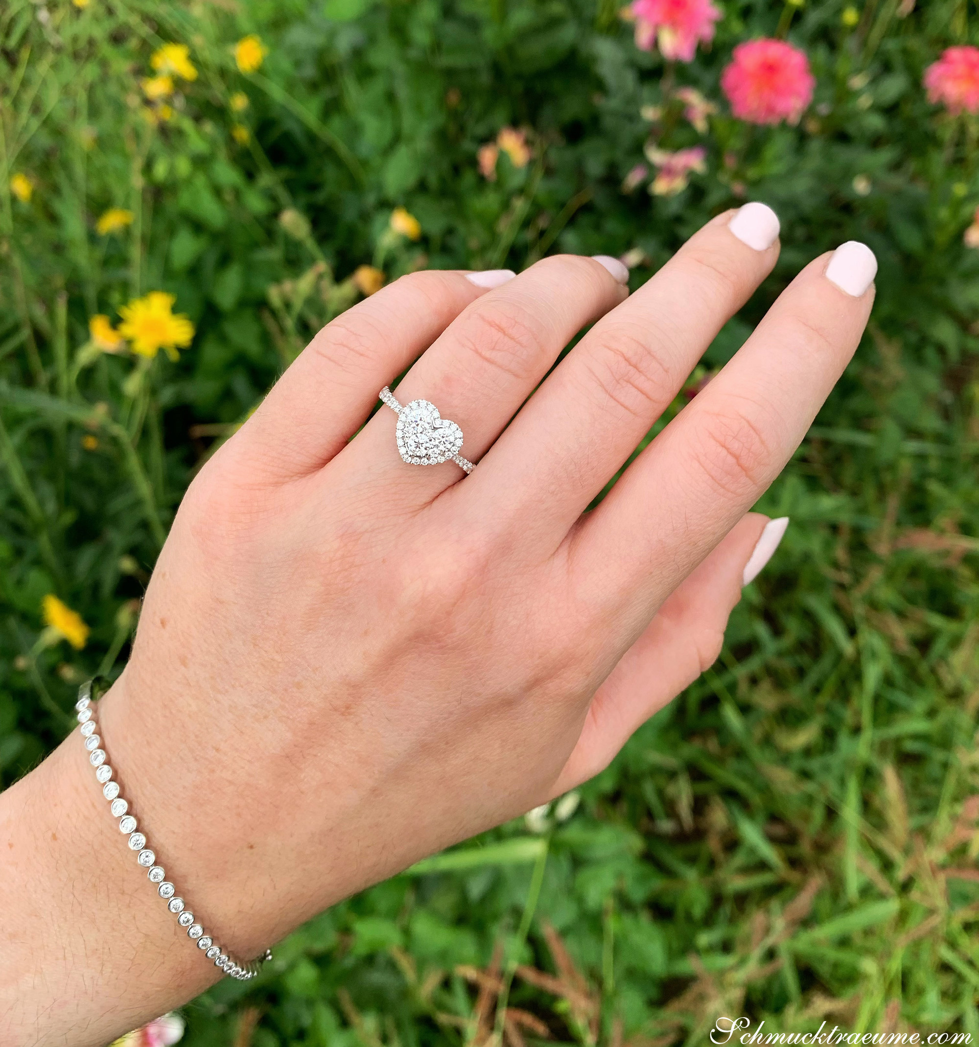 Hand mit Herzform-Brillanten-Ring im Weißgold vor Blumen