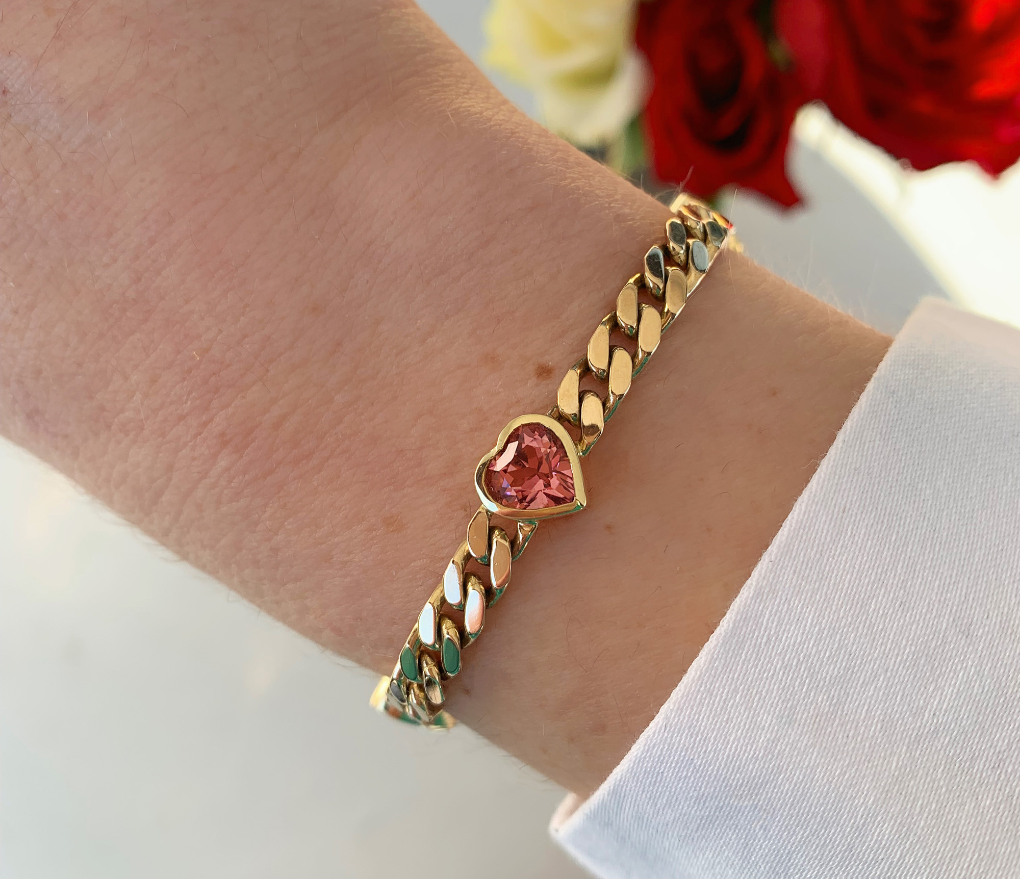 Goldene Panzerarmband mit rosa Turmalin Herz auf Hand mit Rosen im Hintergrund