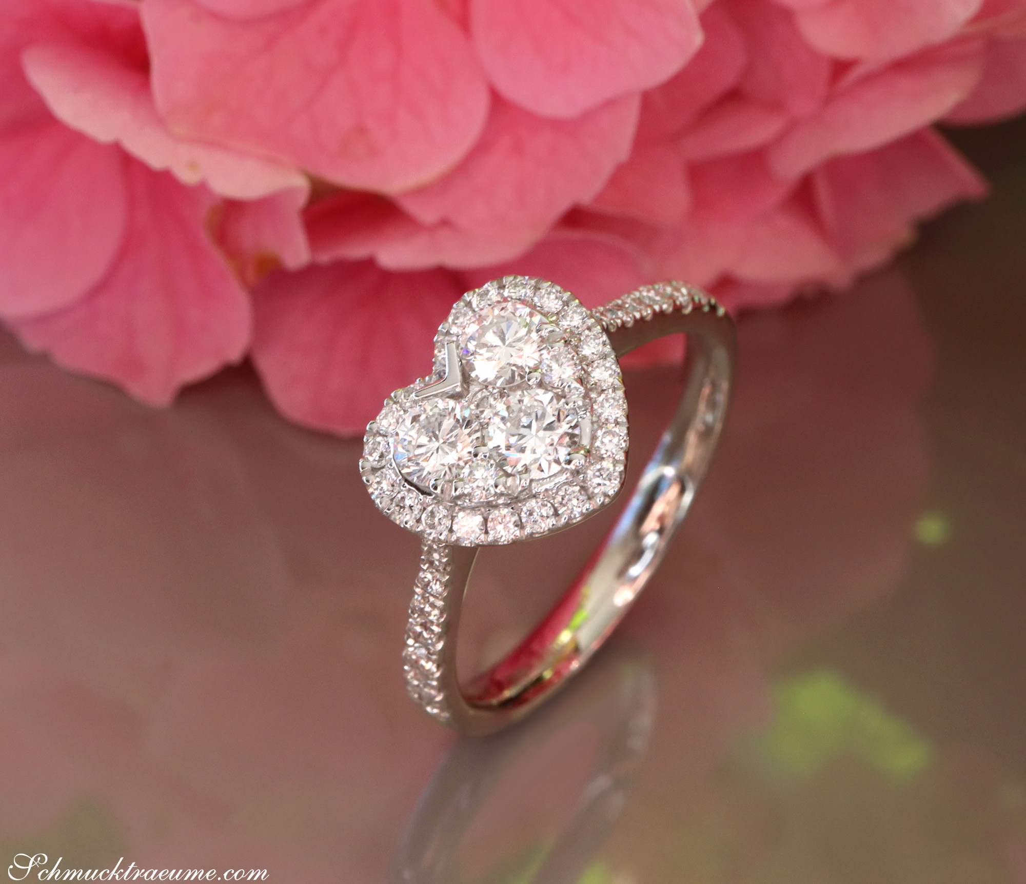 Herzformiger Brillant-Ring mit Diamantbesatz, detailreiches Weißgold, vor rosa Blüten.