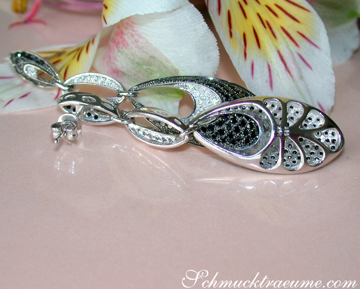 Silberner Ohrhänger mit schwarzen & weißen Diamanten, Detailaufnahme auf rosa Untergrund, Blüten im Hintergrund.