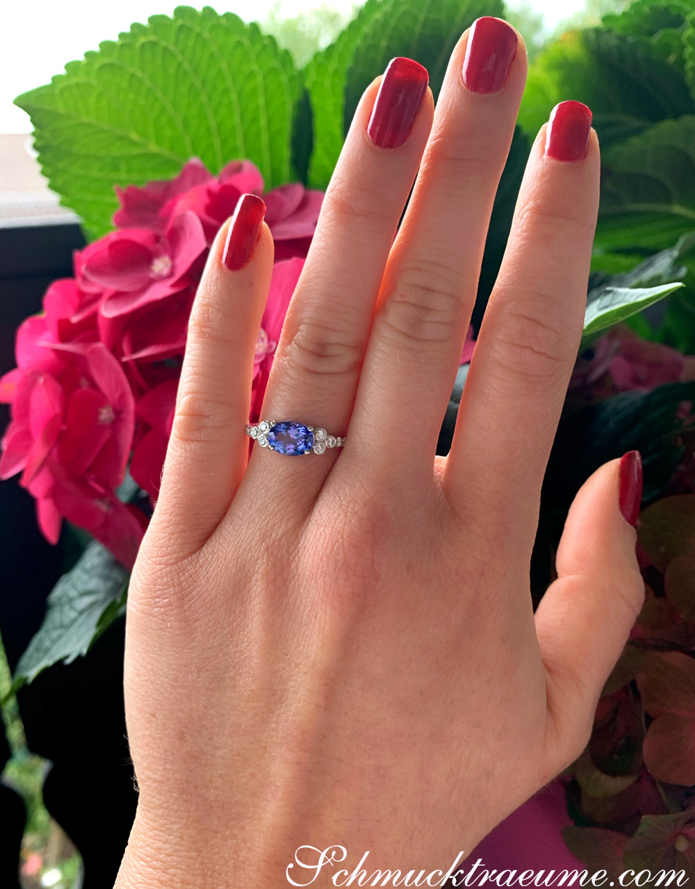 Ringszene mit rotem Nagellack und blauer Steinschmuck vor Blüten