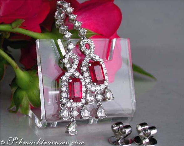 Luxus-Ohrhänger mit Burma-Rubinen und Diamanten, klarer Kristallsockel, rote Rosen im Hintergrund