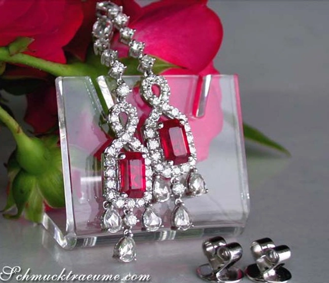 Luxus-Ohrhänger mit Burma-Rubinen und Diamanten, klarer Kristallsockel, rote Rosen im Hintergrund Luxus-Ohrhänger mit Burma-Rubinen und Diamanten, klarer Kristallsockel, rote Rosen im Hintergrund