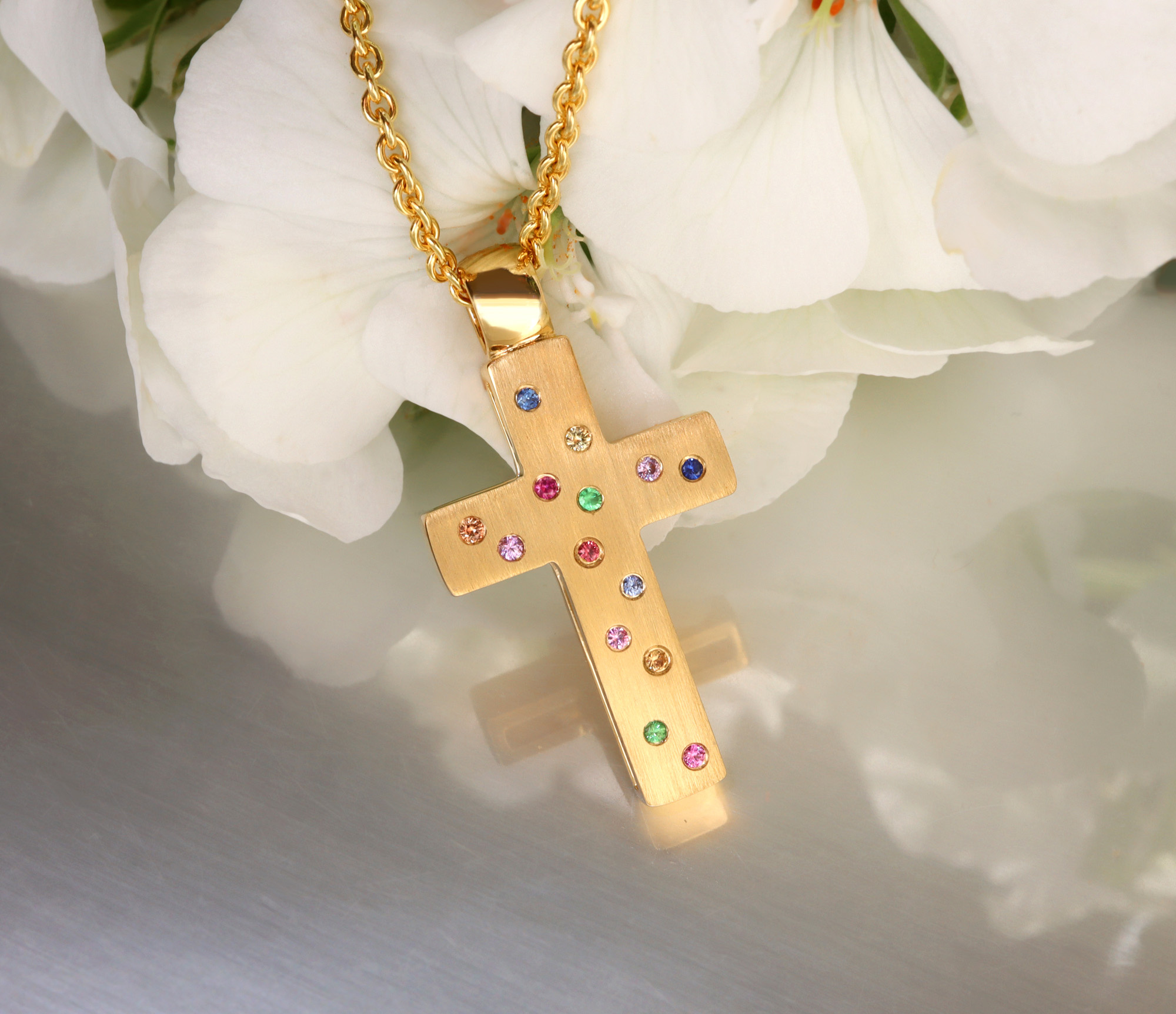 Goldener Kreuzanhänger mit Brillanten und Multicolor Saphiren, Detailaufnahme auf Blütenhintergrund.