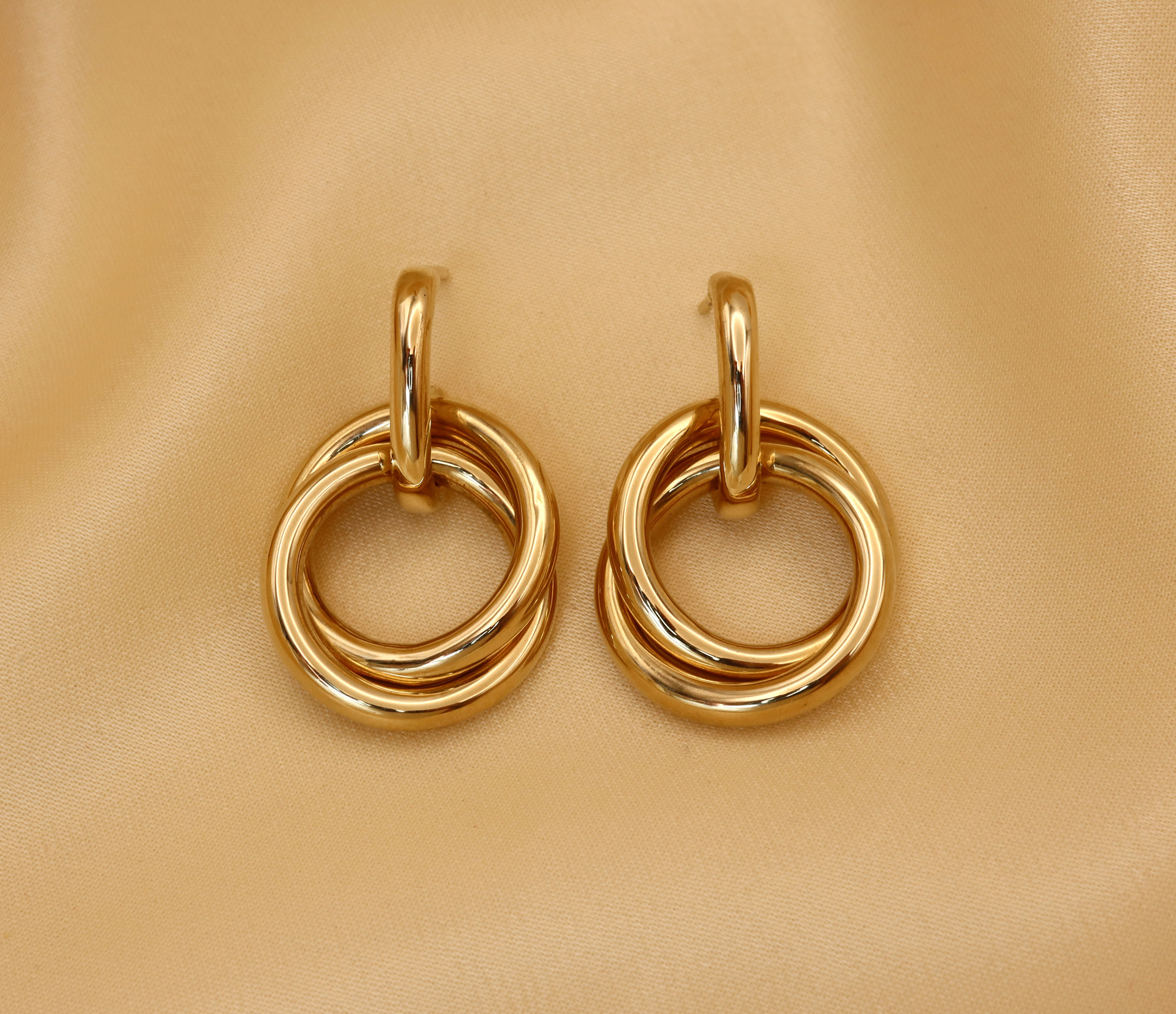 Detailansicht von goldfarbenen Ohrringen mit doppelter geschwungener Ringstruktur auf cremefarbenem Hintergrund.