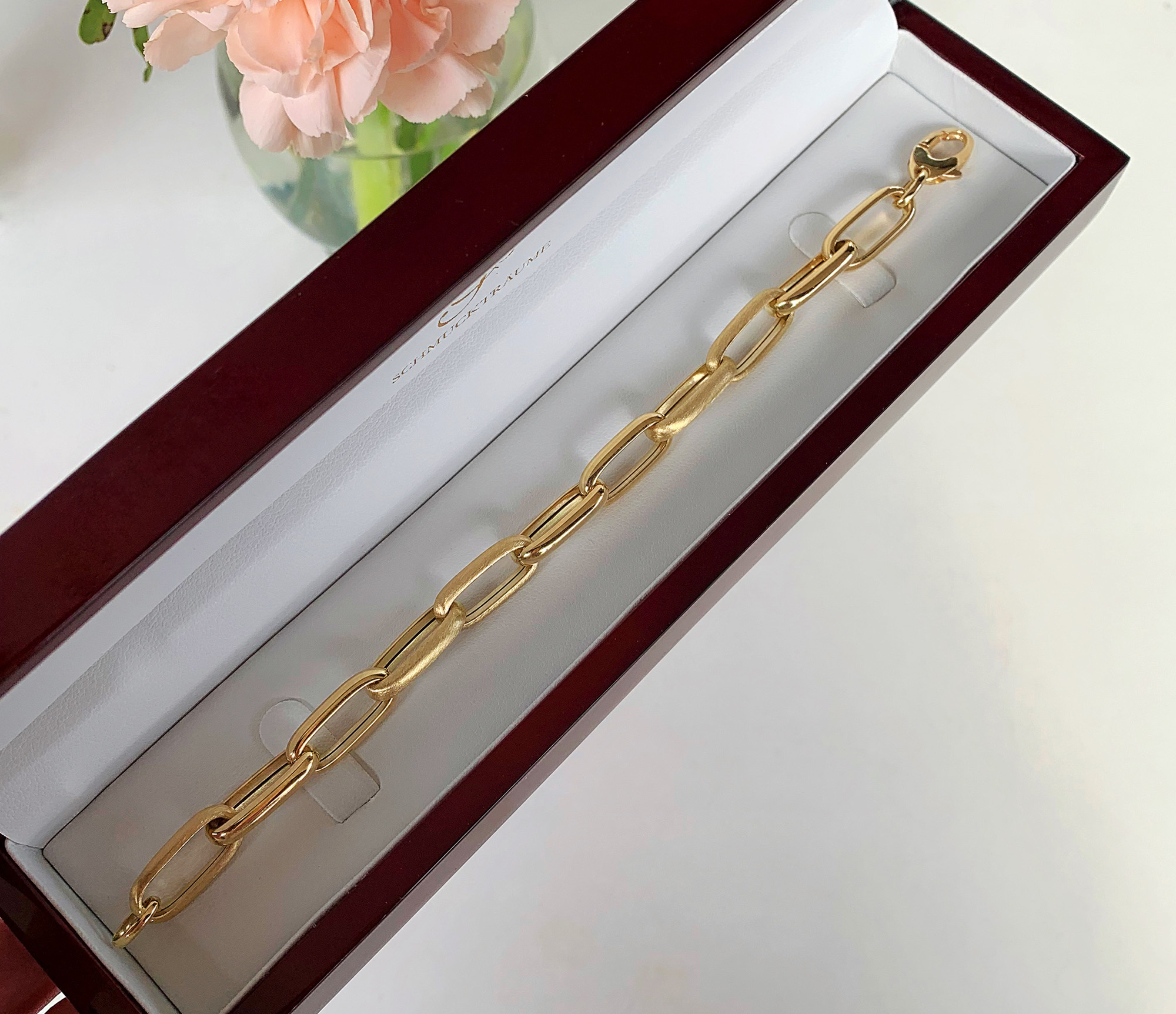 Goldenes Paperclip-Armband in 585er Gelbgold, satiniert und glänzend, in Schmuckbox, Blumen im Hintergrund.