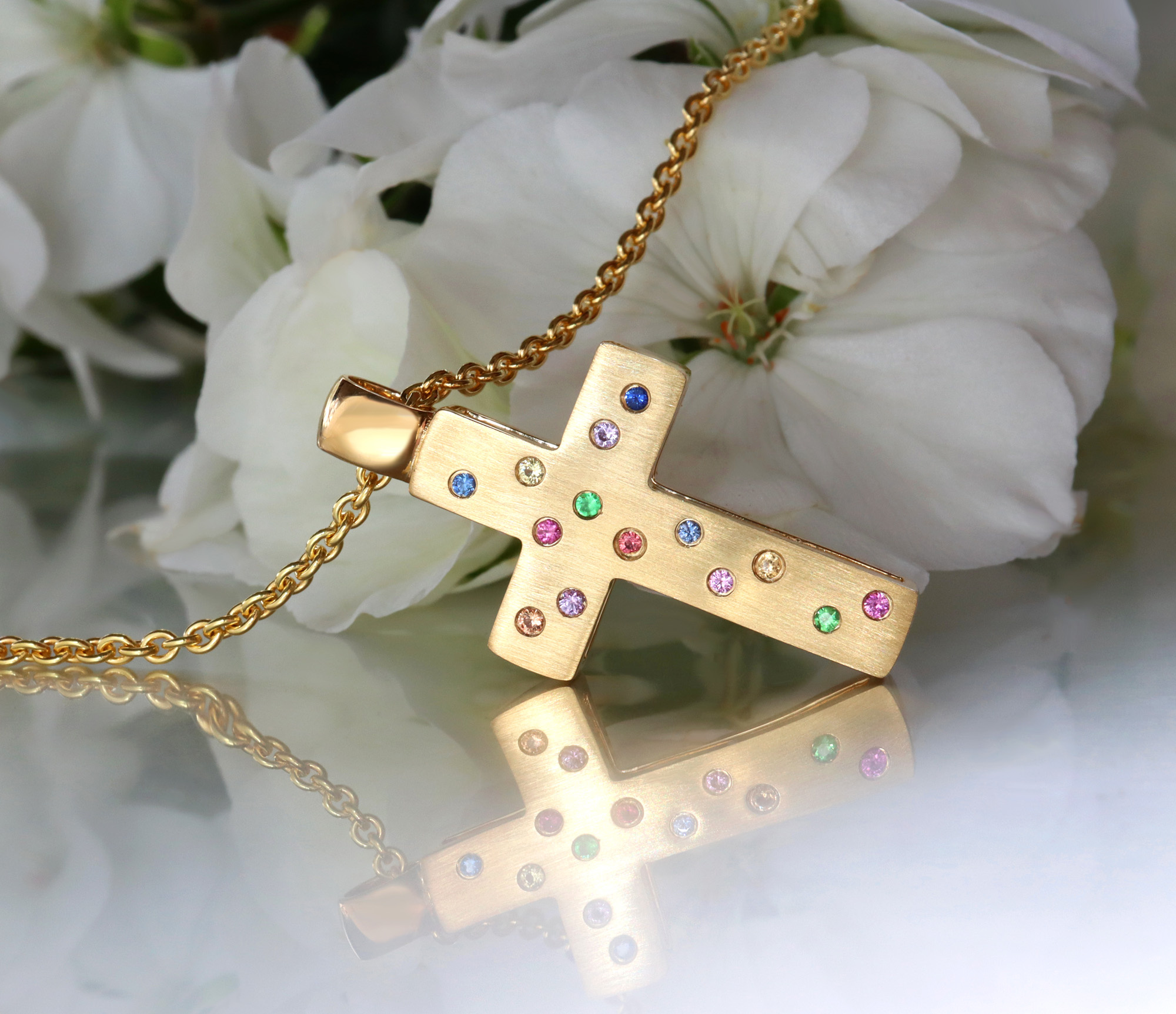 Goldener Kreuzanhänger mit Brillanten und Multicolor Saphiren, Detailaufnahme auf Blütenhintergrund.