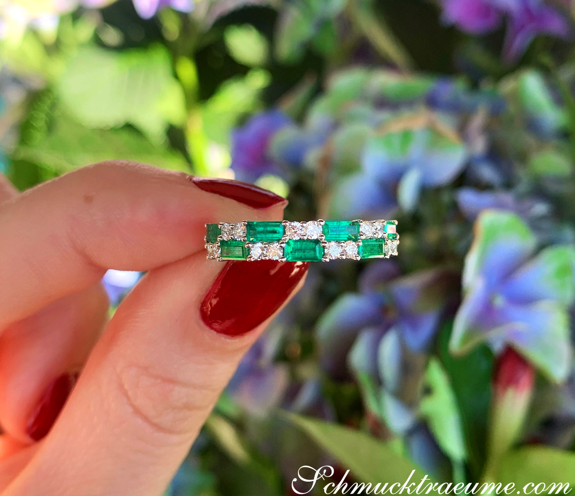 Smaragd- und Brillant-Bandring in Weißgold, Detailaufnahme auf Fingernägel mit rotem Nagellack, Hintergrund mit Blüten.