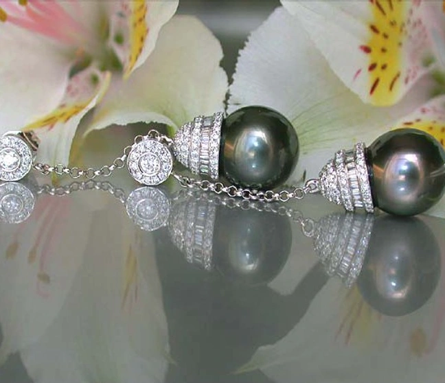 Tahitiperlen-Ohrgehänge mit Brillanten auf reflektierendem Hintergrund, Blüten im Hintergrund. Tahitiperlen-Ohrgehänge mit Brillanten auf reflektierendem Hintergrund, Blüten im Hintergrund.