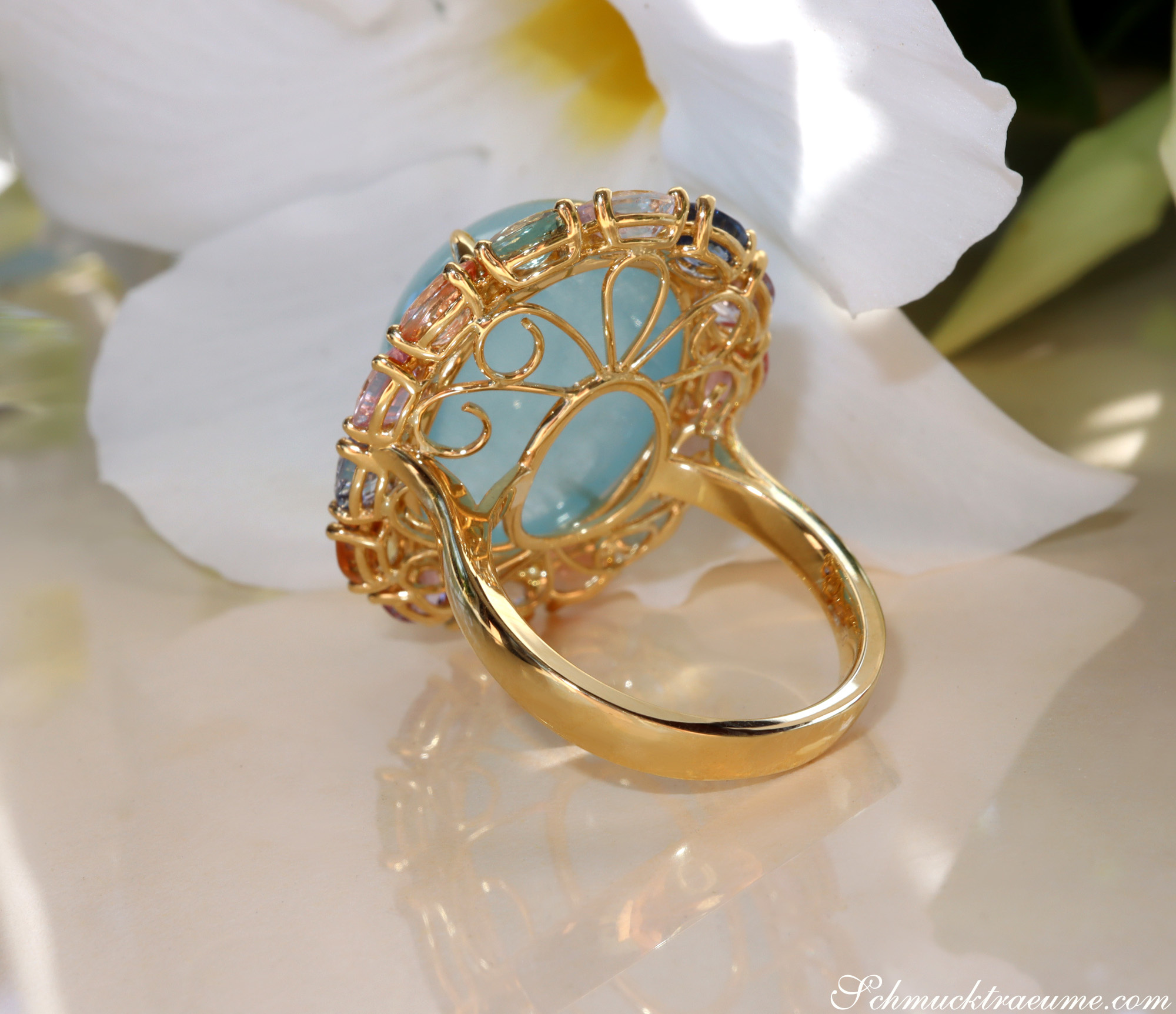 Goldener Ring mit Aquamarin und Saphiren, detailreiche Verzierung, Blüte im Hintergrund.