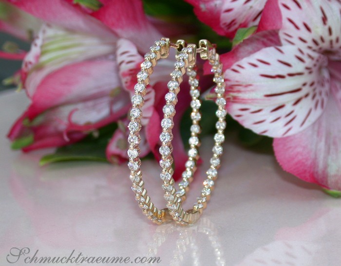 Goldene Creolen mit Diamanten, verziert mit 2.130 Karat, W SI2, auf glänzender Oberfläche, Blumen im Hintergrund.