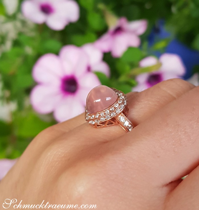 Rosenquarz Herz Ring mit Diamanten in Roségold, Detailaufnahme auf Hand, blühende Blumen im Hintergrund.