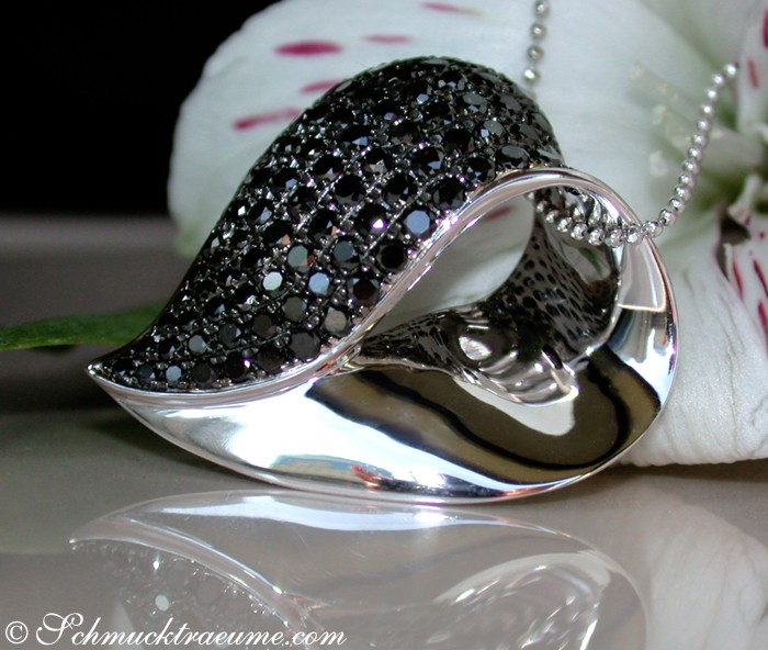 Detailansicht von schwarz-diamantenbesetztem Herzanhänger, glänzende Oberfläche, silberner Hintergrund, weiße Blume im