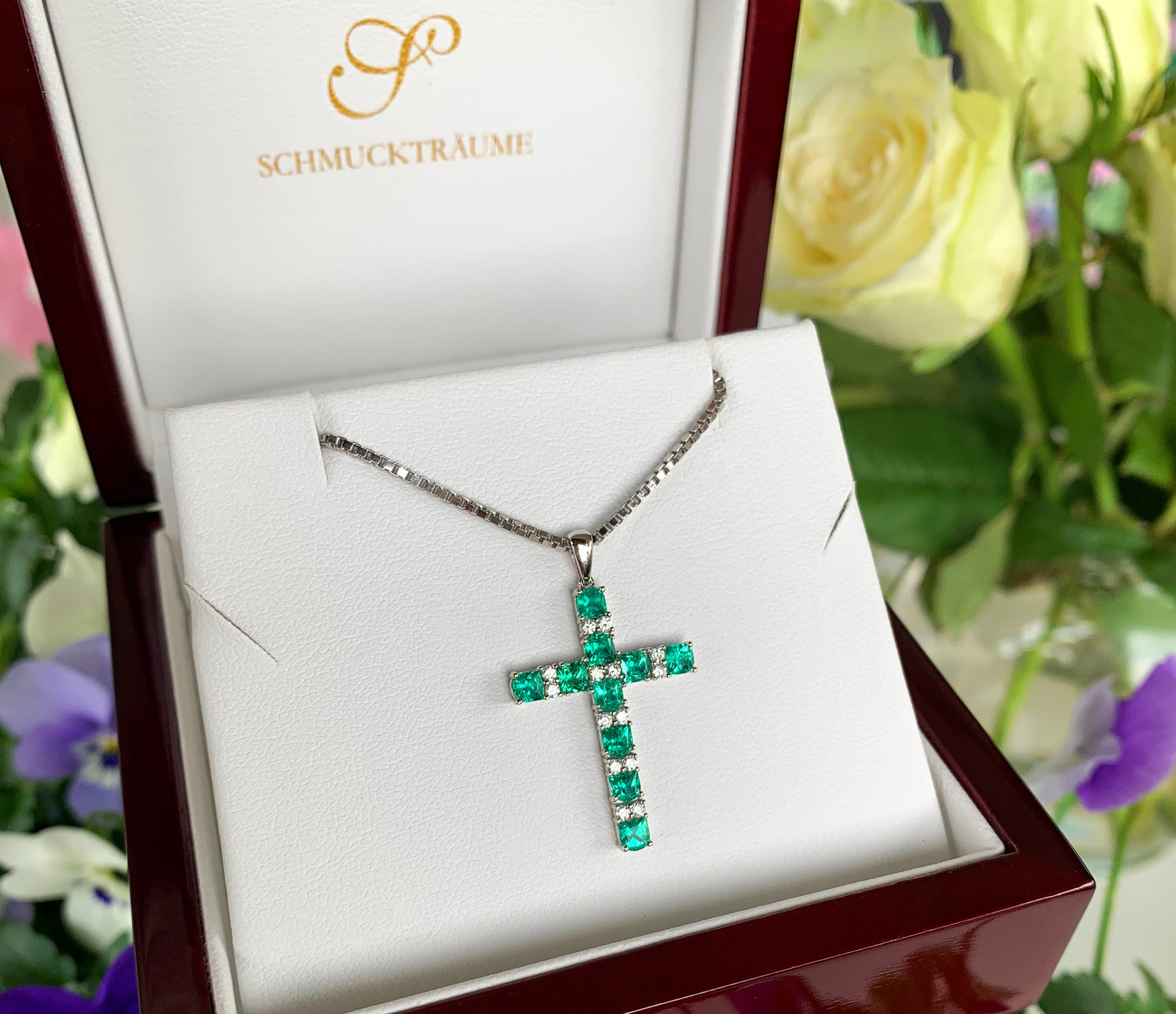 Kreuzanhänger aus Smaragden und Brillanten in Weißgold 750, präsentiert in edler Verpackung mit Rosen im Hintergrund.