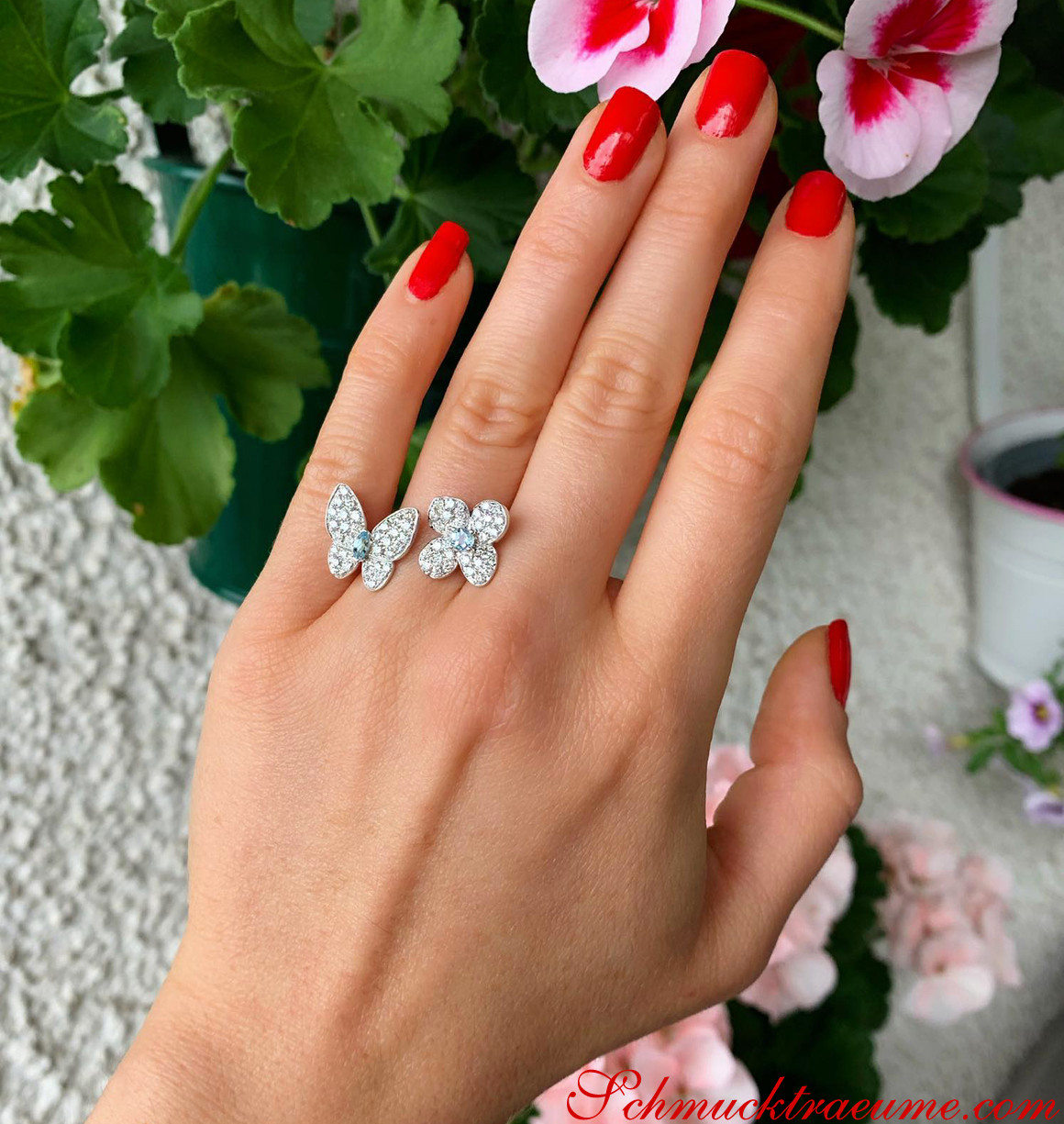 Detailansicht von Damenfinger mit roten Nägeln, elegantem Schmetterlingsring und Blumen im Hintergrund.