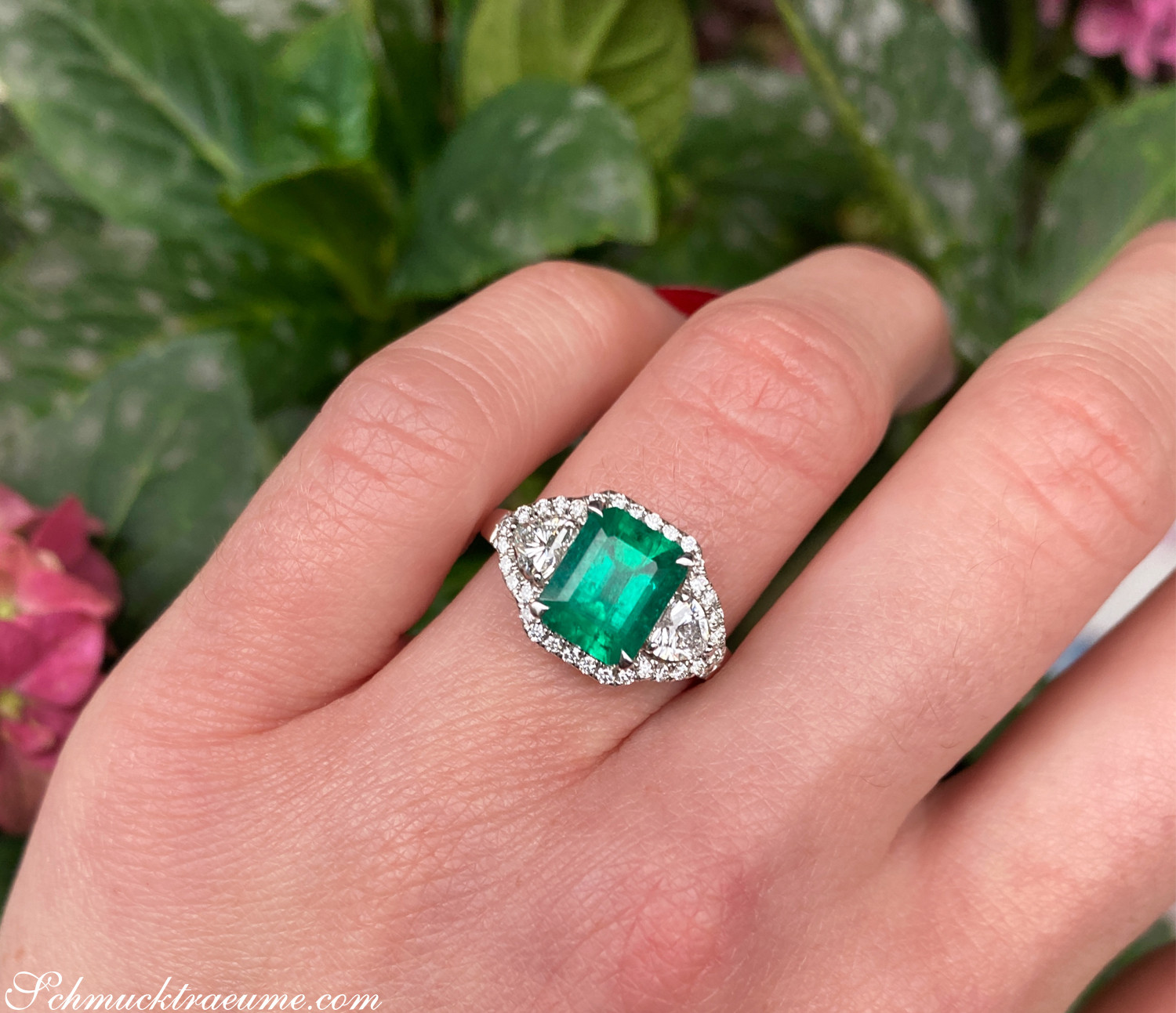 Smaragd-Ring mit Diamant-Herz, detailreich, auf Handfläche getragen, im Freien, vor grünen Blättern.