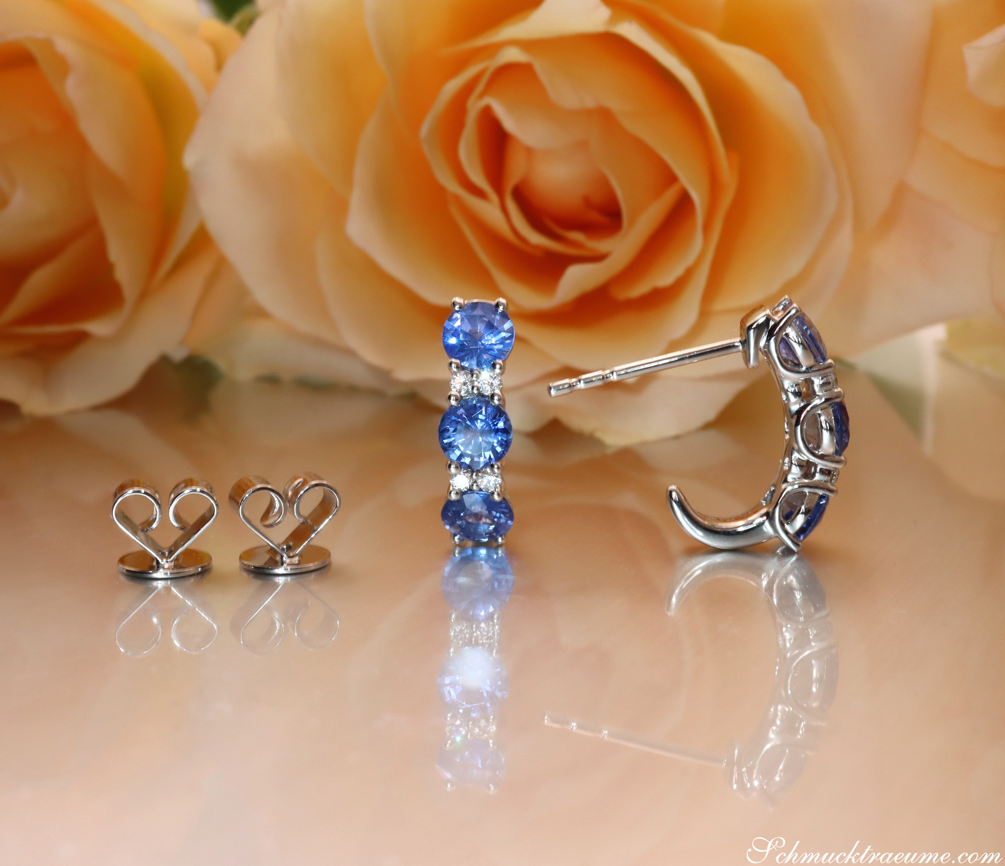 Weißgold-Halbcreolen mit blauen Saphiren und Diamanten, Rosen im Hintergrund.