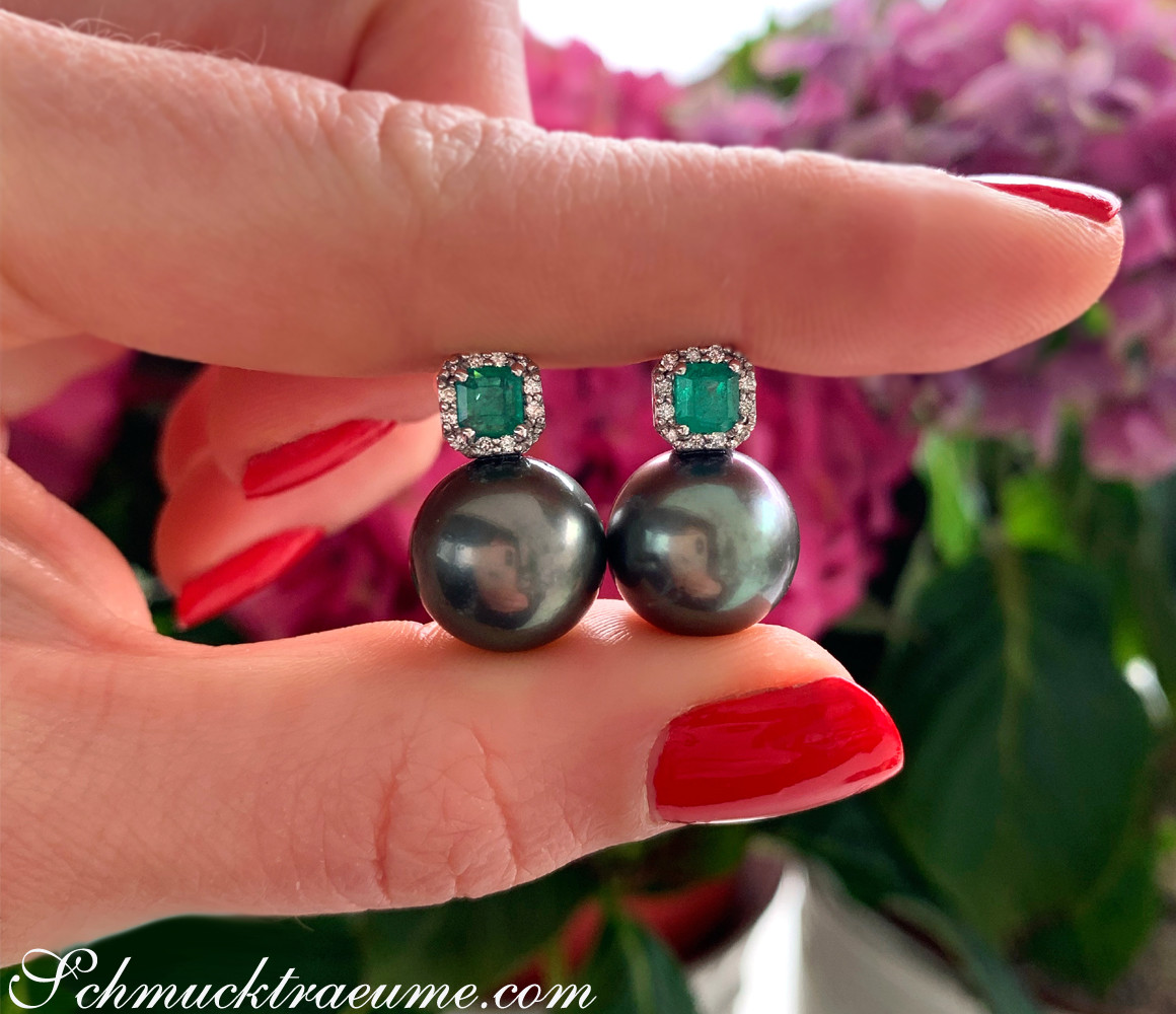 Tahitiperlen-Ohrstecker mit Smaragden und Diamanten, Hand mit rotem Nagellack, Hintergrund mit Blumen.