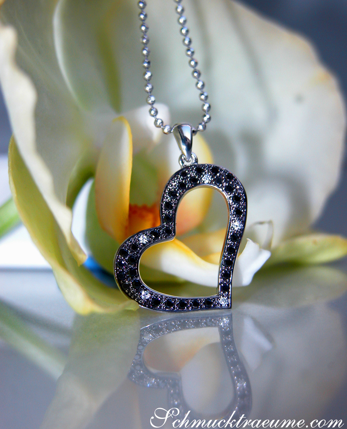 Schwarzer Diamanten Herz-Anhänger auf Kette, zarte Blume im Hintergrund, Detailaufnahme.