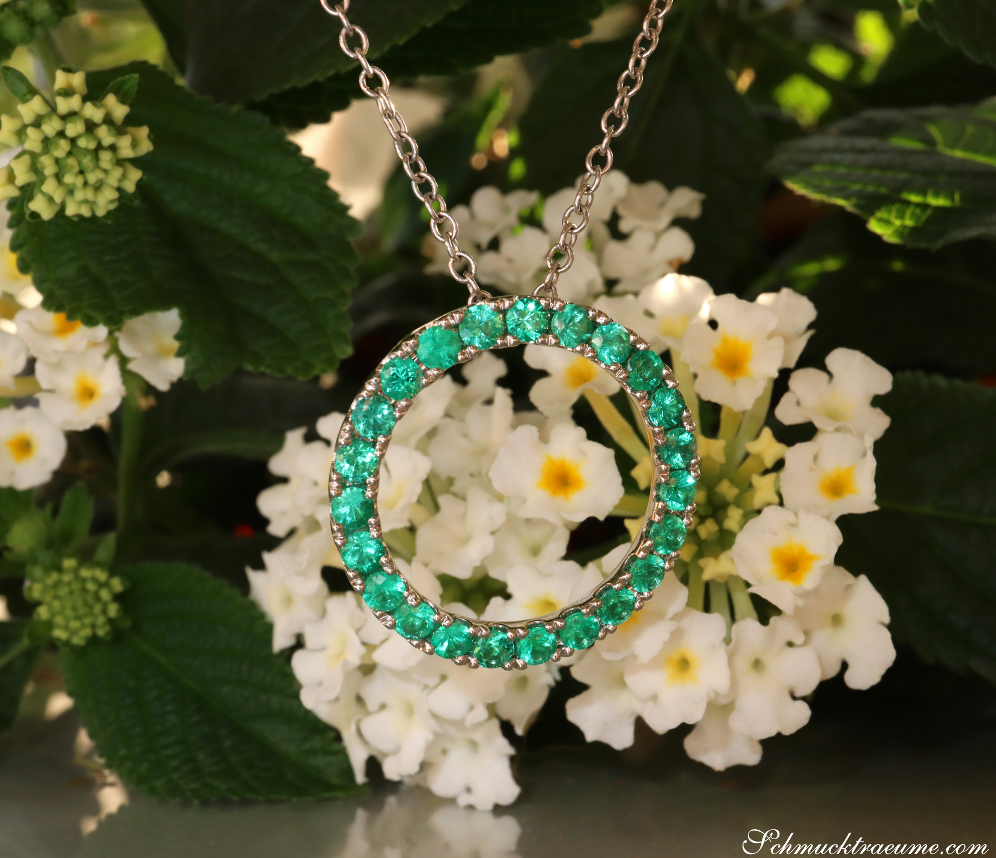 Weißgold-Smaragd-Kreis-Anhänger vor weiß-blühenden Blumen, Detailaufnahme.