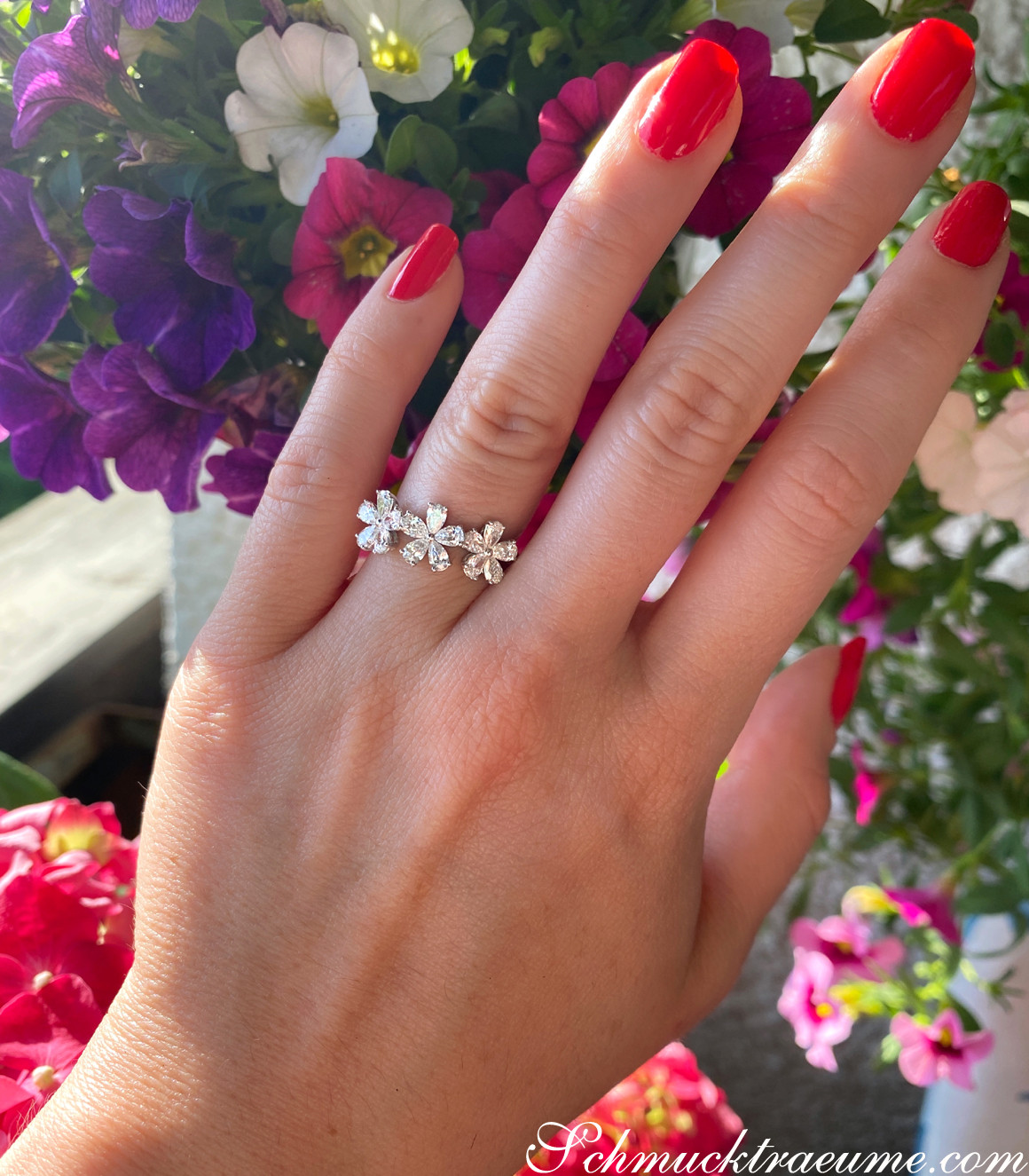 Hand mit roten Nagellack und Diamant-Blüten-Ring vor bunten Blumen