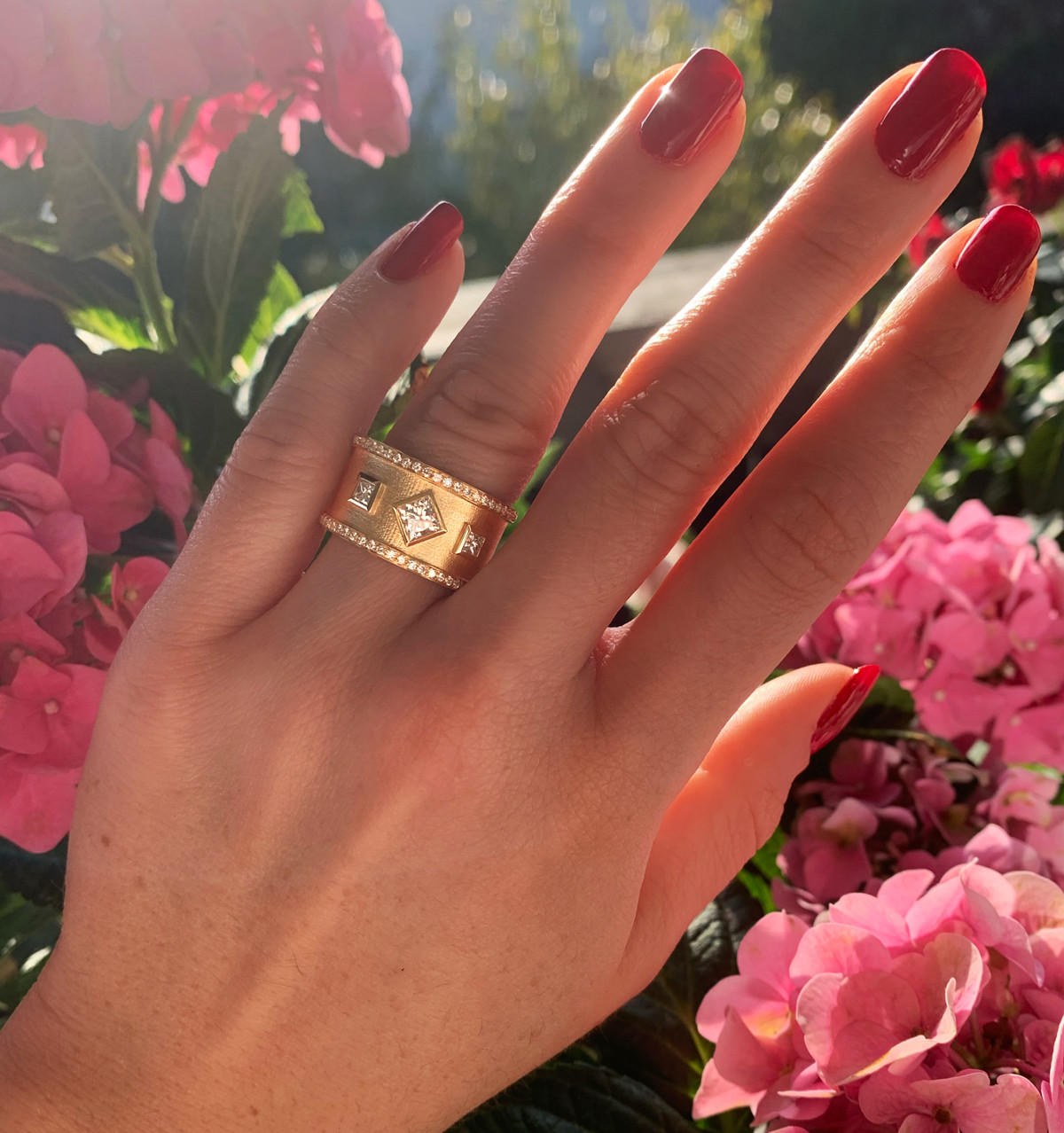 Goldener Prinzess-Diamantring mit drei Diamanten, Nagellack rot, Hintergrund rosa Blüten.