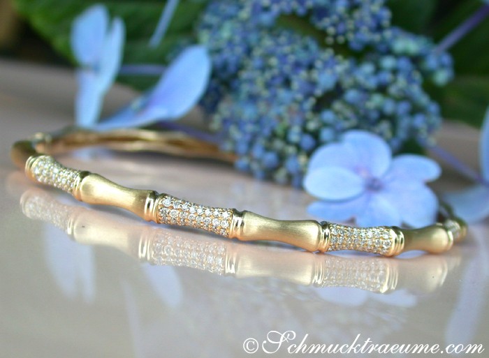 Goldener Bambus-Armreif mit Diamanten auf glänzender Oberfläche, blauer Hortensie im Hintergrund