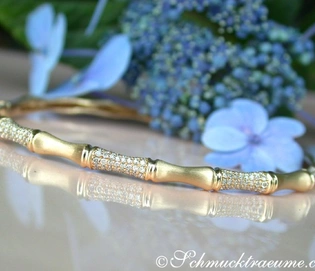 Goldener Bambus-Armreif mit Diamanten auf glänzender Oberfläche, blauer Hortensie im Hintergrund