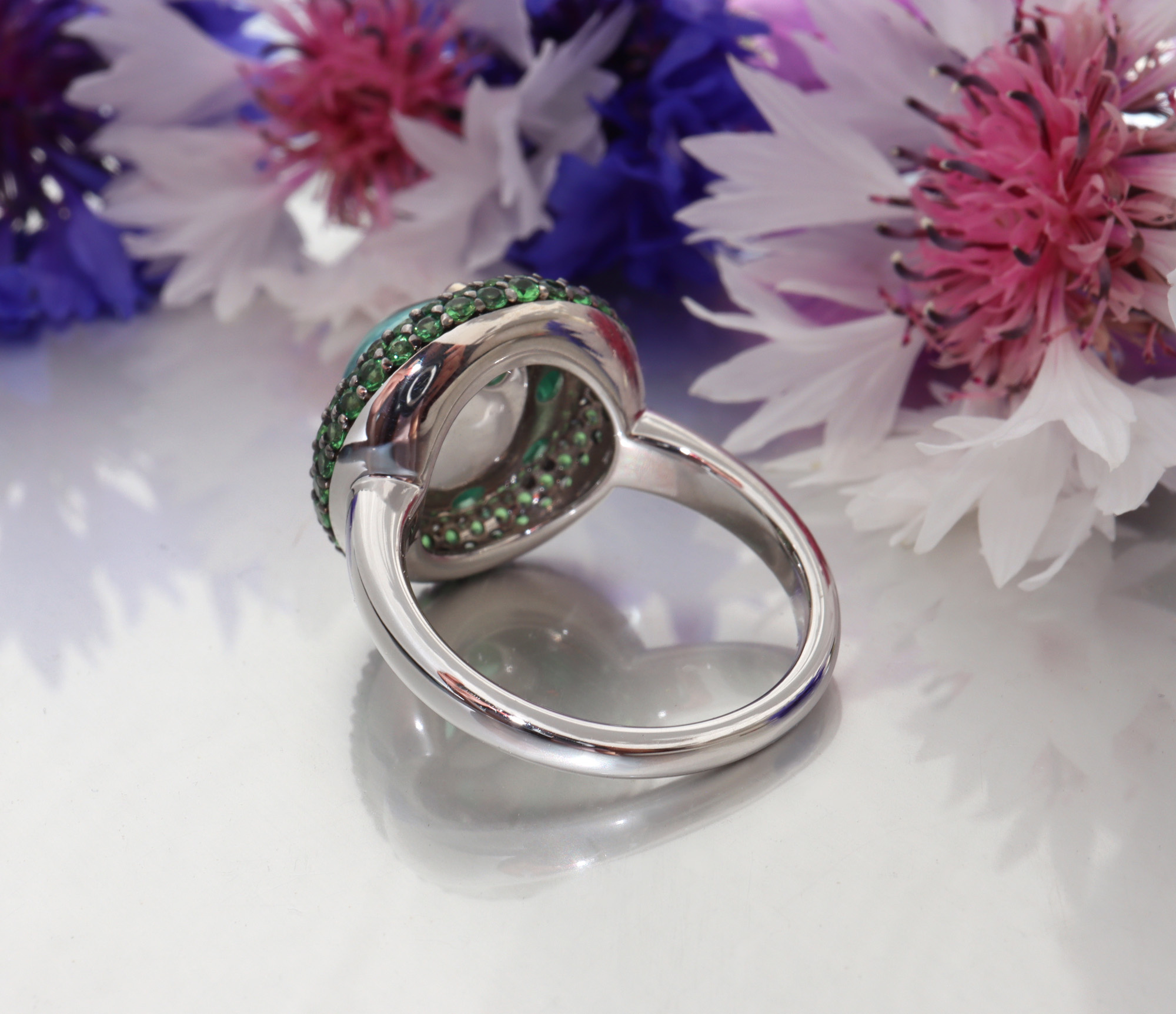 Silberner Ring mit grünen und blauen Steinen, Blumen im Hintergrund