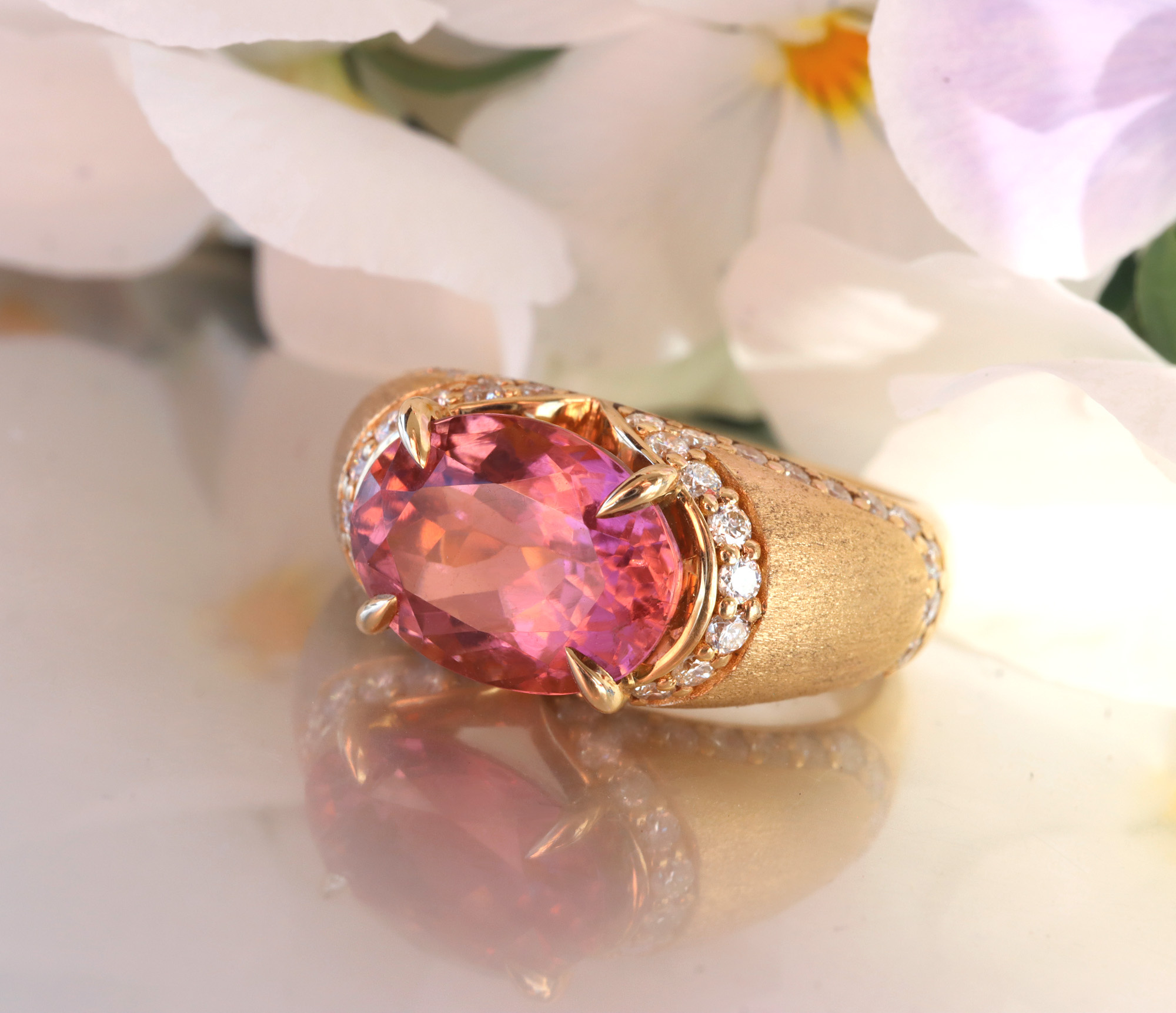 Goldener Ring mit rosa Turmalin und Diamanten, Blumen im Hintergrund.