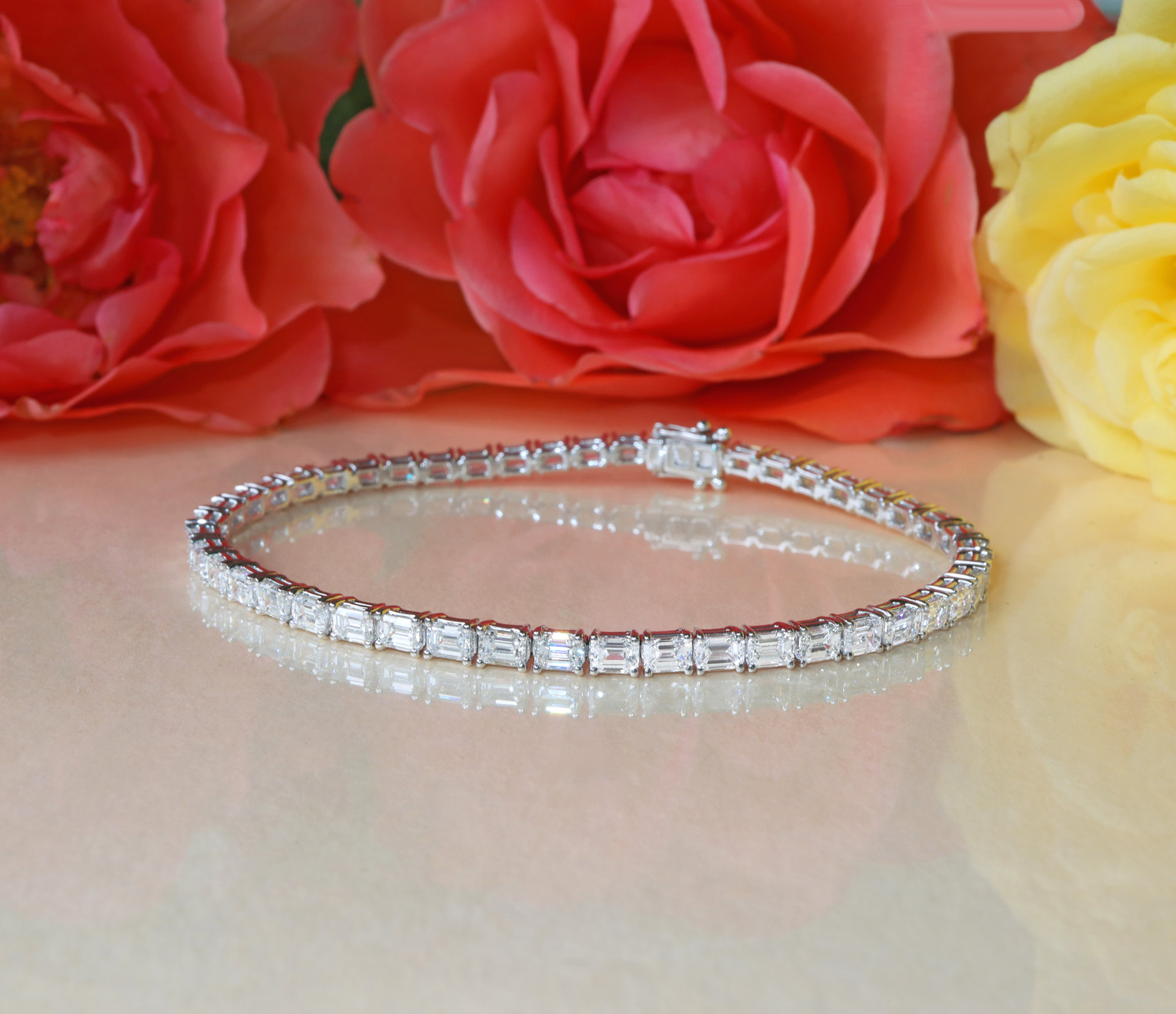 Weißgoldene Tennisarmbänder mit Smaragdschliff-Diamanten auf glänzender Oberfläche, Rosen im Hintergrund.