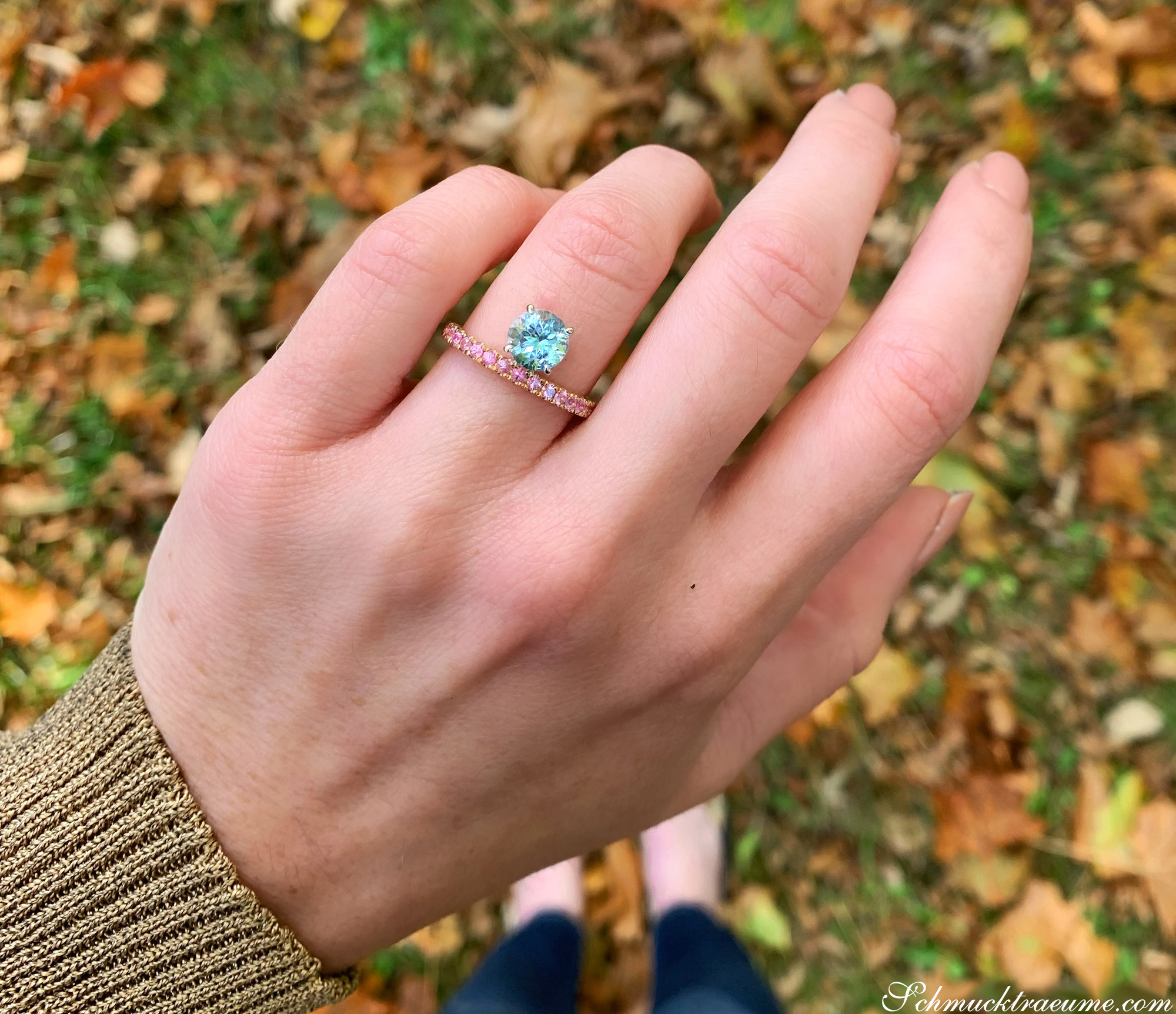 Hand zeigt Aquamarin-Ring mit rosa Saphiren im Freien, Herbstlaub im Hintergrund