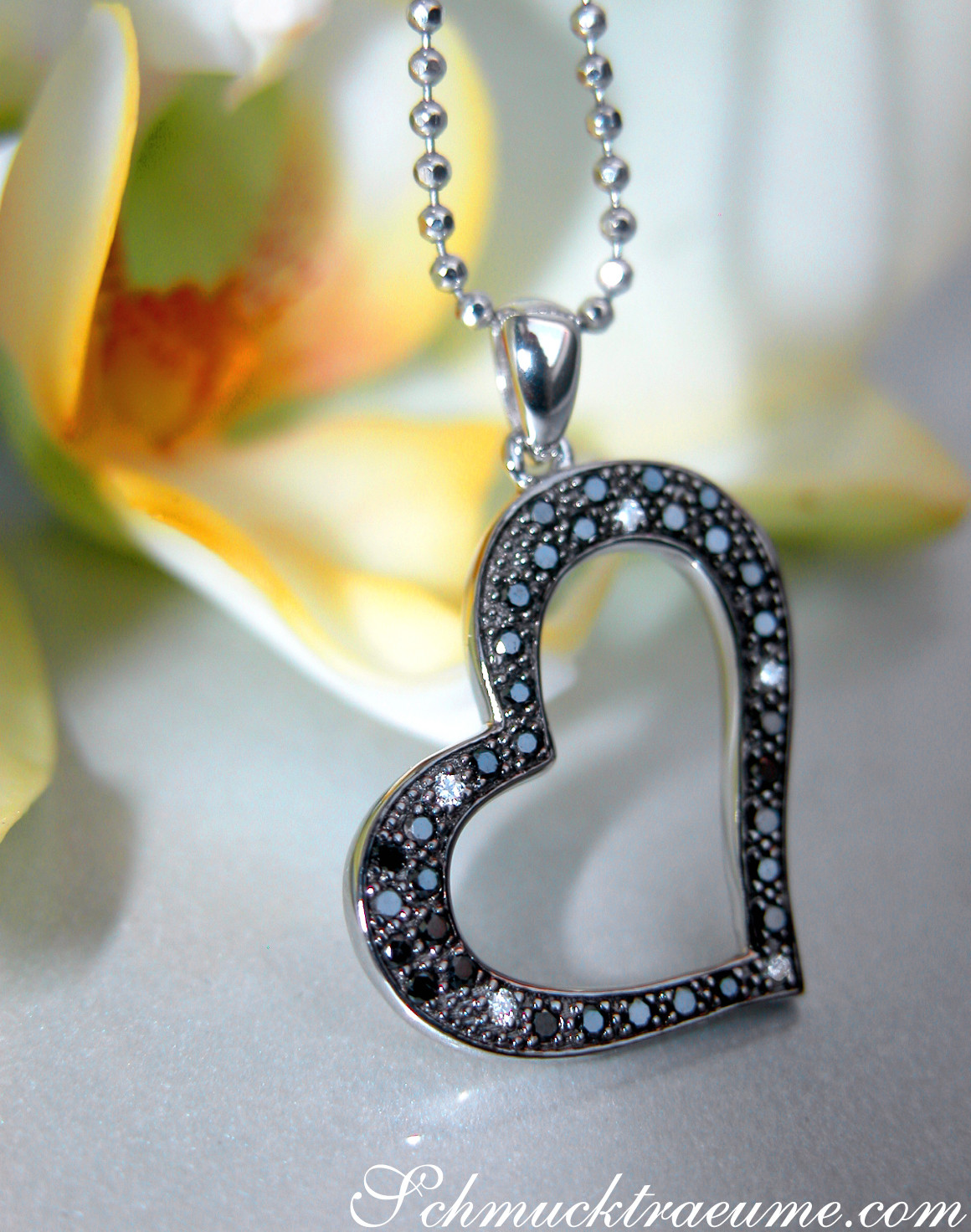 Herz-Anhänger aus Weißgold mit schwarzen Diamanten, detailreich, zarte Kette, Hintergrund Blüte.