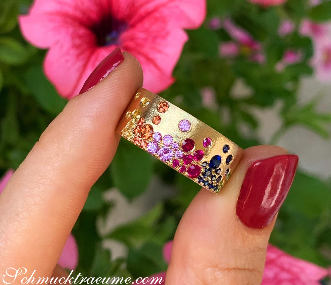 Gelbgoldring mit farbenfrohen Saphiren, Rubinen und Tsavorites, Detailaufnahme, Hintergrund mit Blume