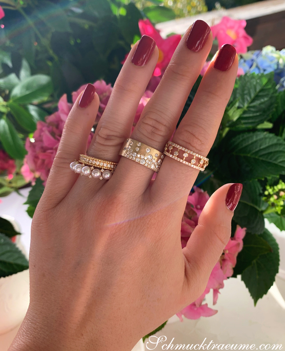 Ringe im Kordel-Design mit Diamanten und Perlen, Nagellack rot, Blumen im Hintergrund