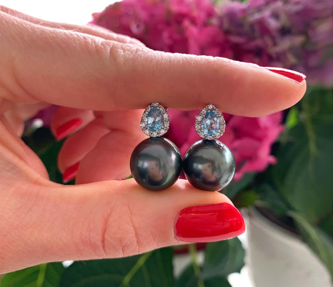 Tahitiperlen-Ohrstecker mit Aquamarinen und Diamanten, Händehalt, rote Nagel, Blüten im Hintergrund.