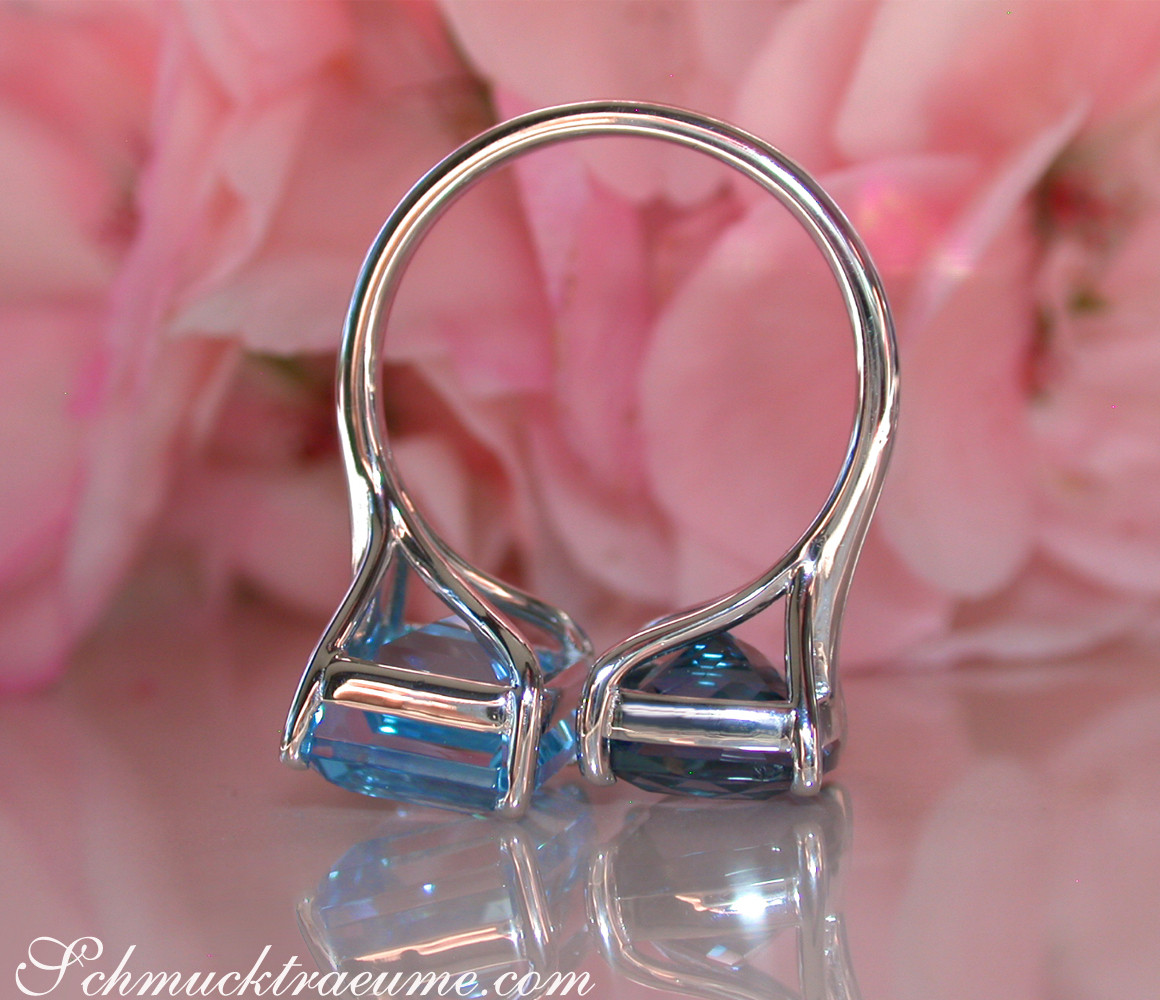 Weißgold-Ring mit Blautopas in eleganten Krappenfassungen, vor Rosenblüten.