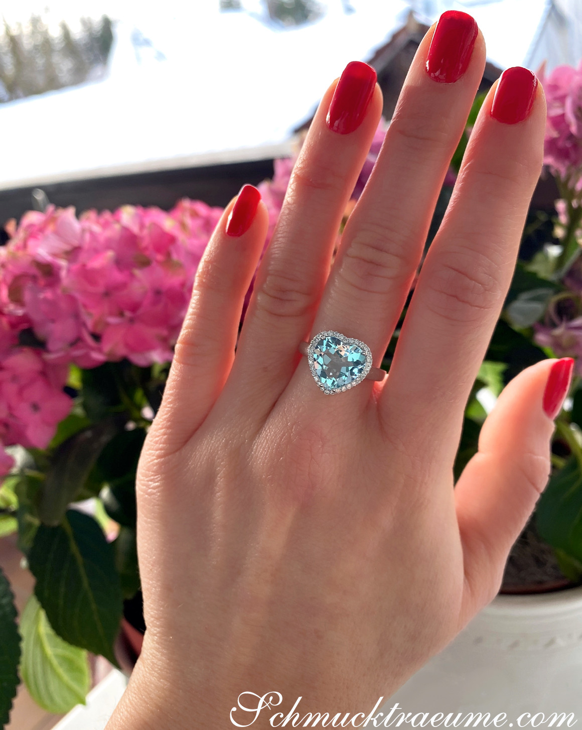 Herzform Aquamarin-Ring mit Diamanten auf Hand mit roten Nagellack vor Blumen